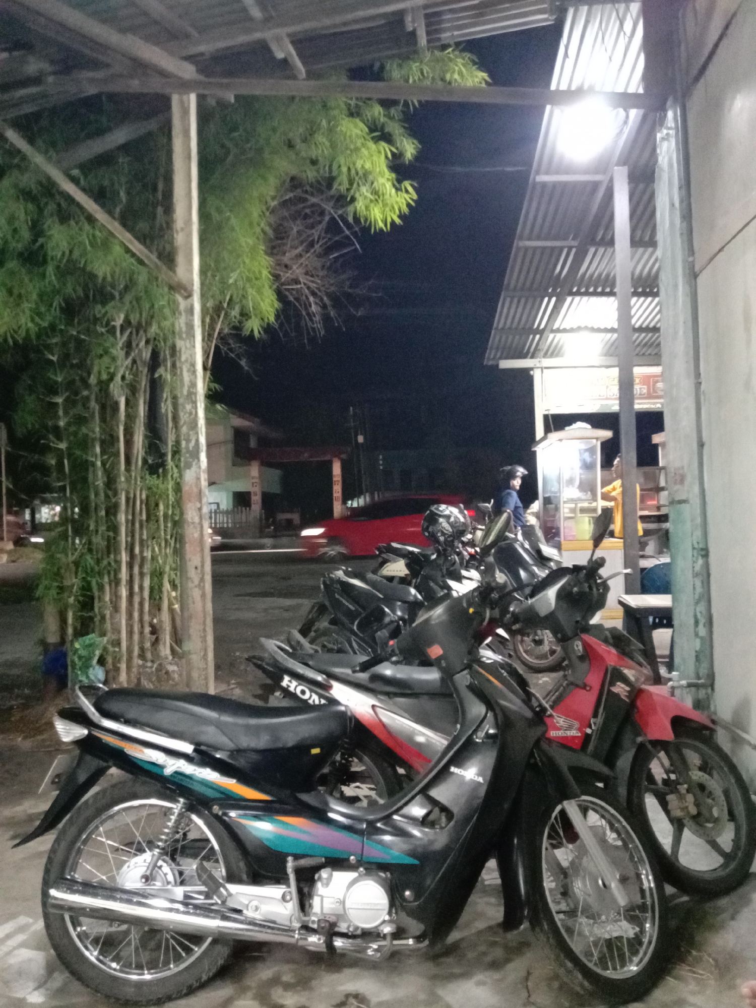 The image shows a street scene in Lhokseumawe, Indonesia, at night. Several motorcycles are parked under a corrugated metal awning supported by concrete pillars. The motorcycles vary in color, including black, red, and black with teal accents, all appearing to be Honda models. In the background, a red car passes by a building with a gated entrance. A food stall or booth is illuminated by a bright light, with two individuals interacting near it. A person wearing a helmet is visible near the stall. Bamboo trees are situated near the left side of the frame. The setting is urban, with a mix of residential and commercial buildings in the background.