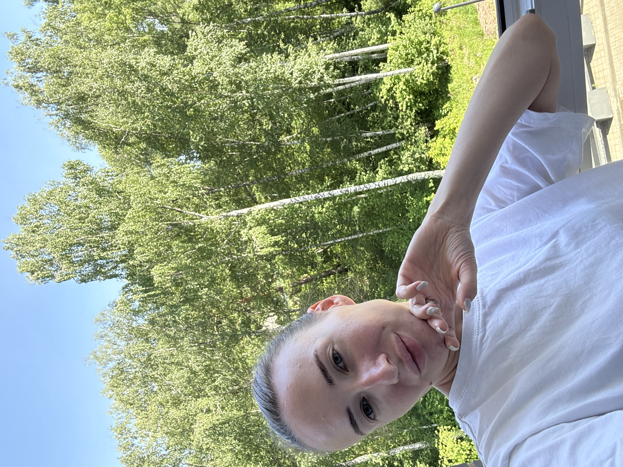 A young Caucasian woman with blonde hair pulled back is taking a selfie outdoors. She is wearing a white t-shirt and her right hand is resting on her chin. The background consists of a dense forest of birch trees under a clear blue sky. The image appears to have been taken on a sunny day.