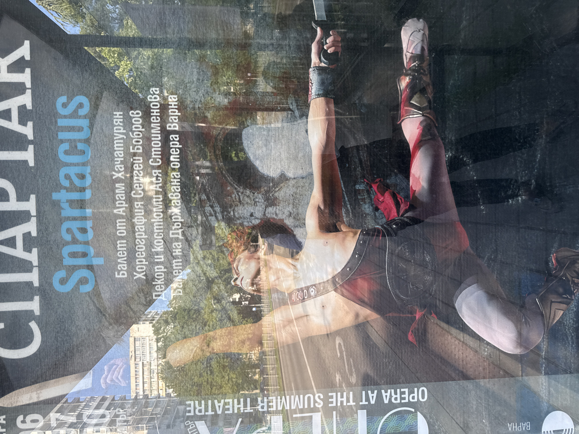 Here's a detailed description of the image:
The image shows a poster for the ballet "Spartacus" by Aram Khachaturian, with choreography by Sergei Bobrov and decor and costumes by Asya Stoimenova. The poster is advertising a performance by the State Opera of Varna. The central figure on the poster is a male dancer, dressed in a theatrical gladiator costume. He is holding a knife upwards.
The poster is mounted on a surface. There are reflections on the surface, including a street scene with a bridge and vegetation. Additionally, there is a partial reflection of a building that looks like a theater. In the lower portion of the poster is "OPERA AT THE SUMMER THEATRE." It also indicates that the opera is in Varna.