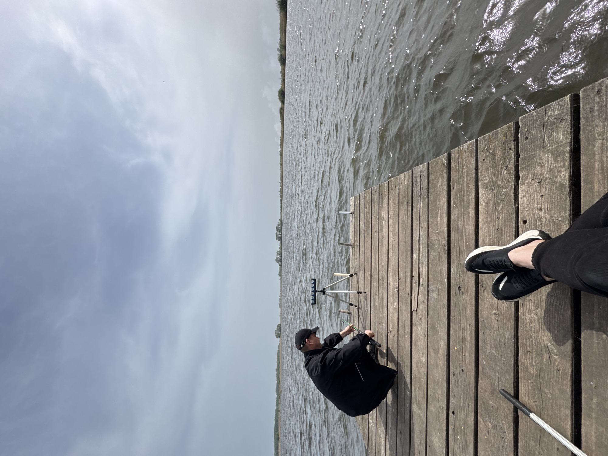 A view from a wooden dock depicts a man in a black jacket and baseball cap preparing fishing rods by a tripod rod stand. The sky is overcast. The water of the lake is choppy, and vegetation lines the distant shore. Beside the man, another person's legs are visible on the dock. A white and black walking stick lies nearby. The location is Unknown, Unknown.