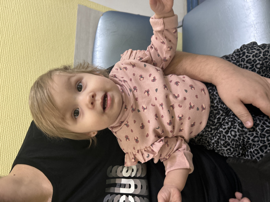 The image captures a young child seated on an adult's lap within an indoor environment in the city of Chaikovsky, Russia. The specific internal location is not identifiable. The child, appearing to be an infant or toddler, has light brown hair, fair skin, and dark eyes, looking directly forward with a partially open mouth revealing lower teeth. Small gold stud earrings are present. She is dressed in a ribbed, light pink long-sleeved turtleneck shirt with ruffled shoulders and a pattern of small, dark red/brown, and grey abstract shapes. Her lower body is clad in dark grey and black leopard-print leggings. The adult, whose chest, arms, and hands are visible, wears a black t-shirt with a layered white "FOCUS" graphic across the chest, supporting the child. The background consists of a textured, light yellow wall. To the right of the child, a light blue institutional-style chair with a metallic frame is visible, along with a portion of a light-colored horizontal surface above it.