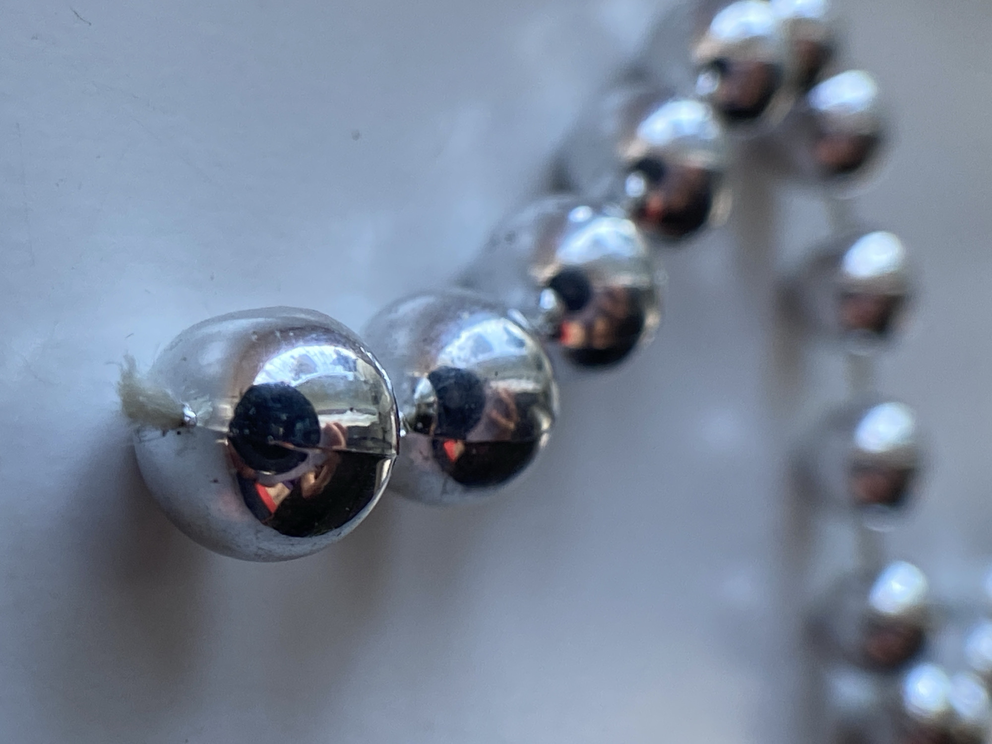 The image shows a close-up, shallow depth of field view of a string of small, silver, spherical beads. The beads are arranged in a line, receding into the background. The surface of the beads is highly reflective, showing distorted reflections of what appears to be a person and possibly the surrounding environment, including a warm red light. The beads are strung together with a thin white cord, visible at the bottom of the closest bead. The background is a plain, light-colored surface, likely a table or countertop. The lighting suggests an indoor setting, possibly illuminated by natural light and artificial light sources. No specific location in a city is identifiable.