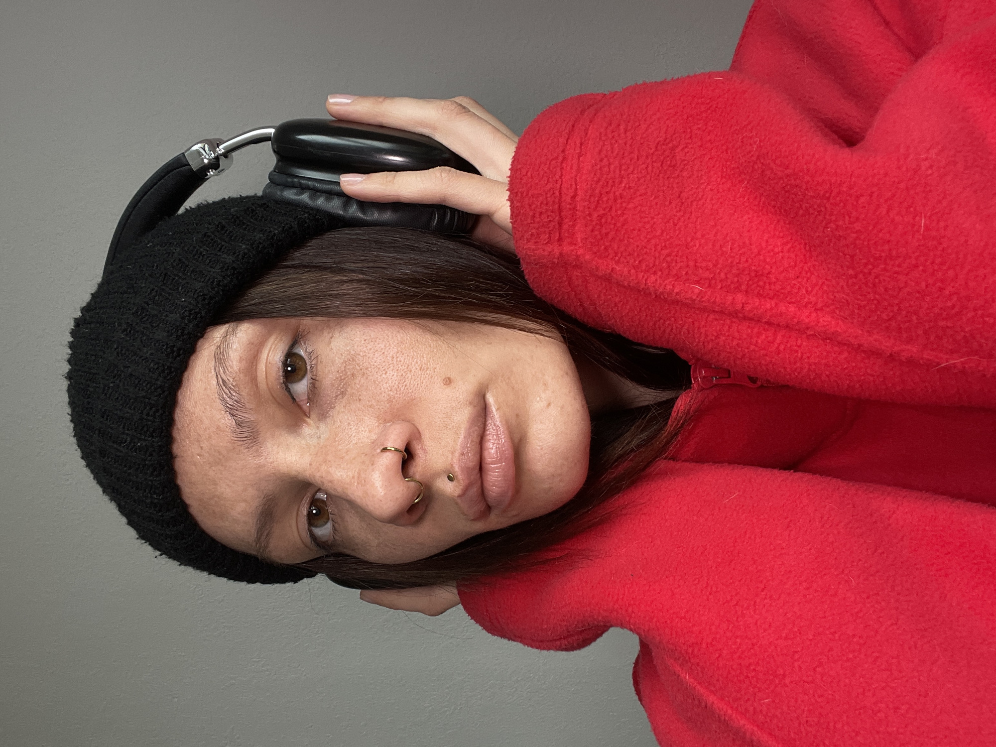 A close-up, portrait-style photograph depicts a young woman looking directly at the viewer. She has dark brown hair, visible below a black knit beanie, and warm brown eyes. Her skin features numerous freckles across her nose and cheeks. Notable facial piercings include a gold ring in her left nostril, a gold hoop through her septum, and a gold stud as a labret piercing below her lower lip.
She is wearing a bright red fleece jacket or hoodie. Her right hand is positioned near her temple, holding a pair of black over-ear headphones with metallic silver accents against her head. The background is a plain, light grey wall, suggesting an indoor setting with even lighting. The woman maintains a neutral to slightly serious expression. No text is visible in the image.