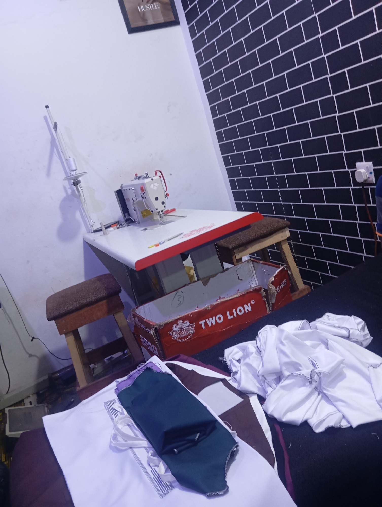 A sewing machine is set up on a table in a room. The table is white with red trim and has a sewing machine on it. There are two stools at the table. In the foreground, there are some pieces of fabric. In the background, there is a wall with black and white brick pattern and a picture frame on the wall. The room is dimly lit.
