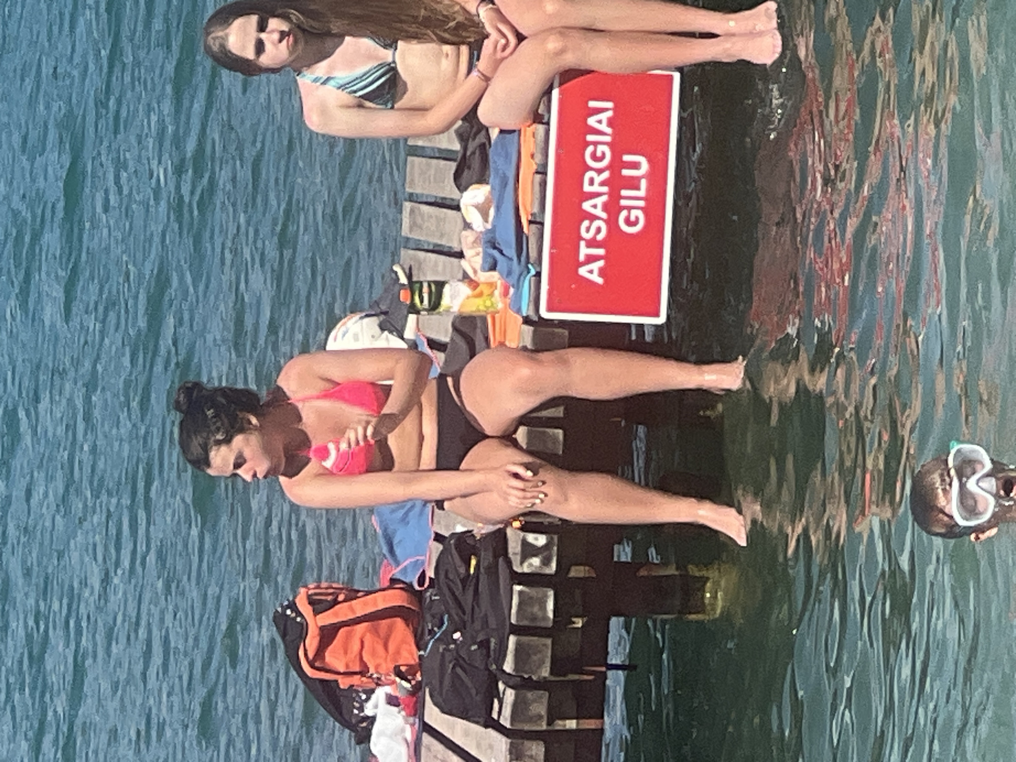 The image shows two young women sitting on a wooden pier next to a body of water, likely a lake or river, on a sunny day. Both women are wearing swimwear. The woman on the left is wearing a striped bikini and has long, light brown hair. The woman on the right is wearing a pink bikini top and black bottoms and has her dark hair pulled up in a bun. She is holding a pink towel in her hands. Behind them, a red sign with white text is visible. The text on the sign reads "ATSARGIAI GILU". In the water to the right, a person wearing a snorkel mask is visible. The water appears calm and reflects the surroundings. Several bags, including an orange and black duffel bag and a black backpack, are placed on the pier. The overall scene suggests a recreational activity, possibly swimming or sunbathing, in a natural setting near Vilnius, Lithuania. The bright sunlight and clear water indicate pleasant weather conditions.