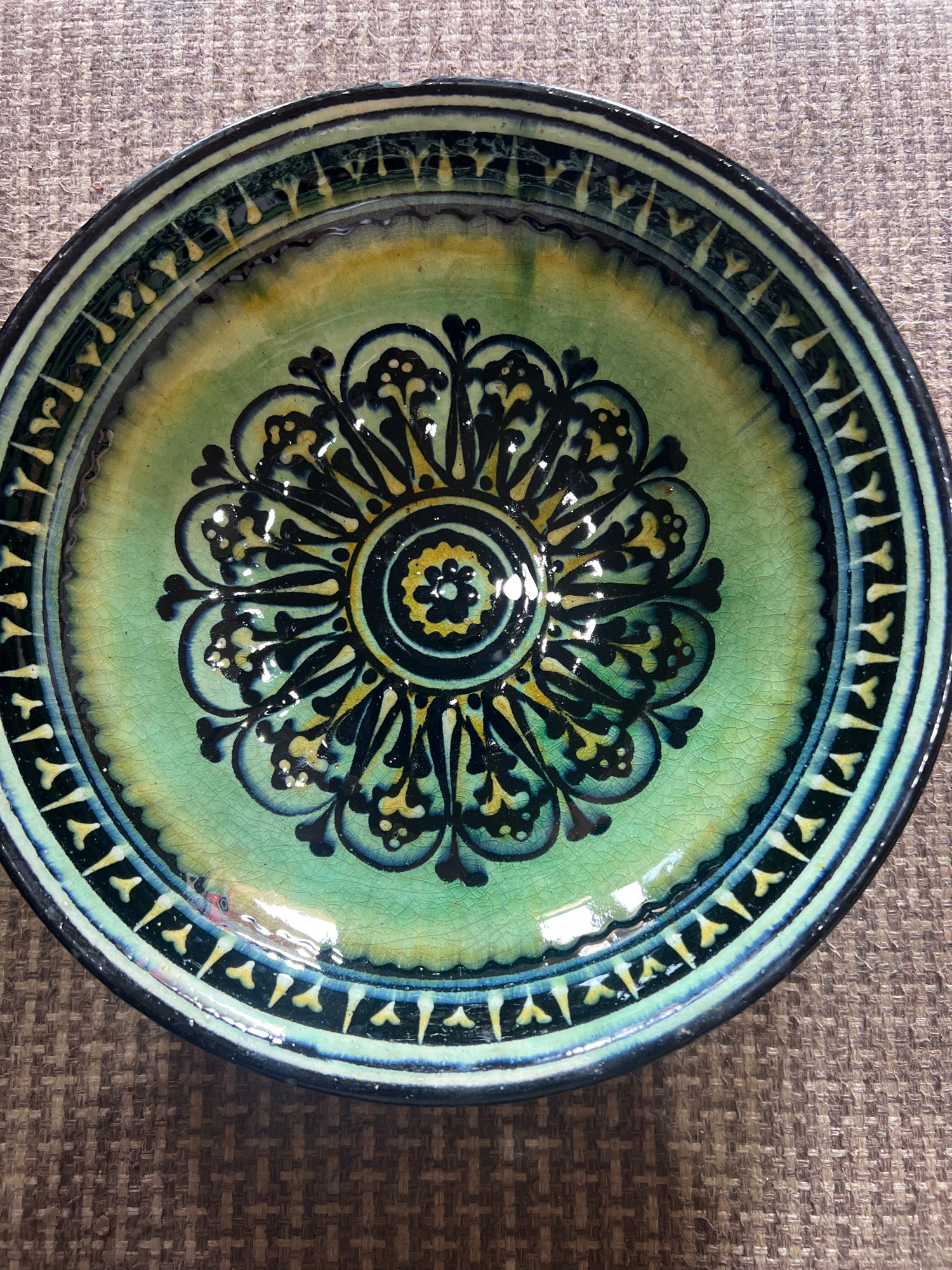 This is a top-down, close-up, forensic shot of a decorative ceramic bowl. The bowl is circular and features intricate black and yellow floral patterns on a green glazed background. The outer rim of the bowl has a decorative band of alternating black and light blue segments with yellow accents. The bowl rests on a textured, brown surface, possibly a tablecloth or placemat. The image does not contain any information that would suggest a specific location in Karamanas, Uzbekistan.