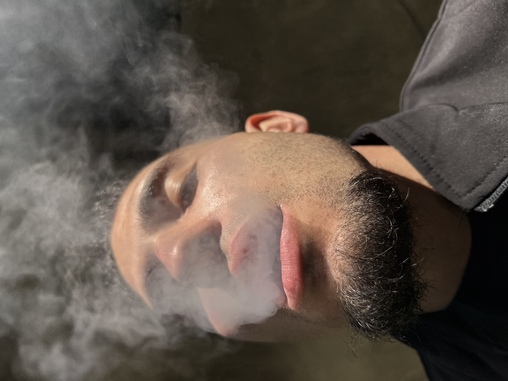 An eye-level, close-up photograph shows a person, seemingly male, exhaling white smoke or vapor. The person has short, dark hair, visible facial hair, and light skin. The background is dark and indistinct. The individual is wearing a dark, possibly black, garment. The image does not reveal any information about the location in Shumen, Bulgaria. There is no information about the action or interaction. The environment is only dark or indistinct.