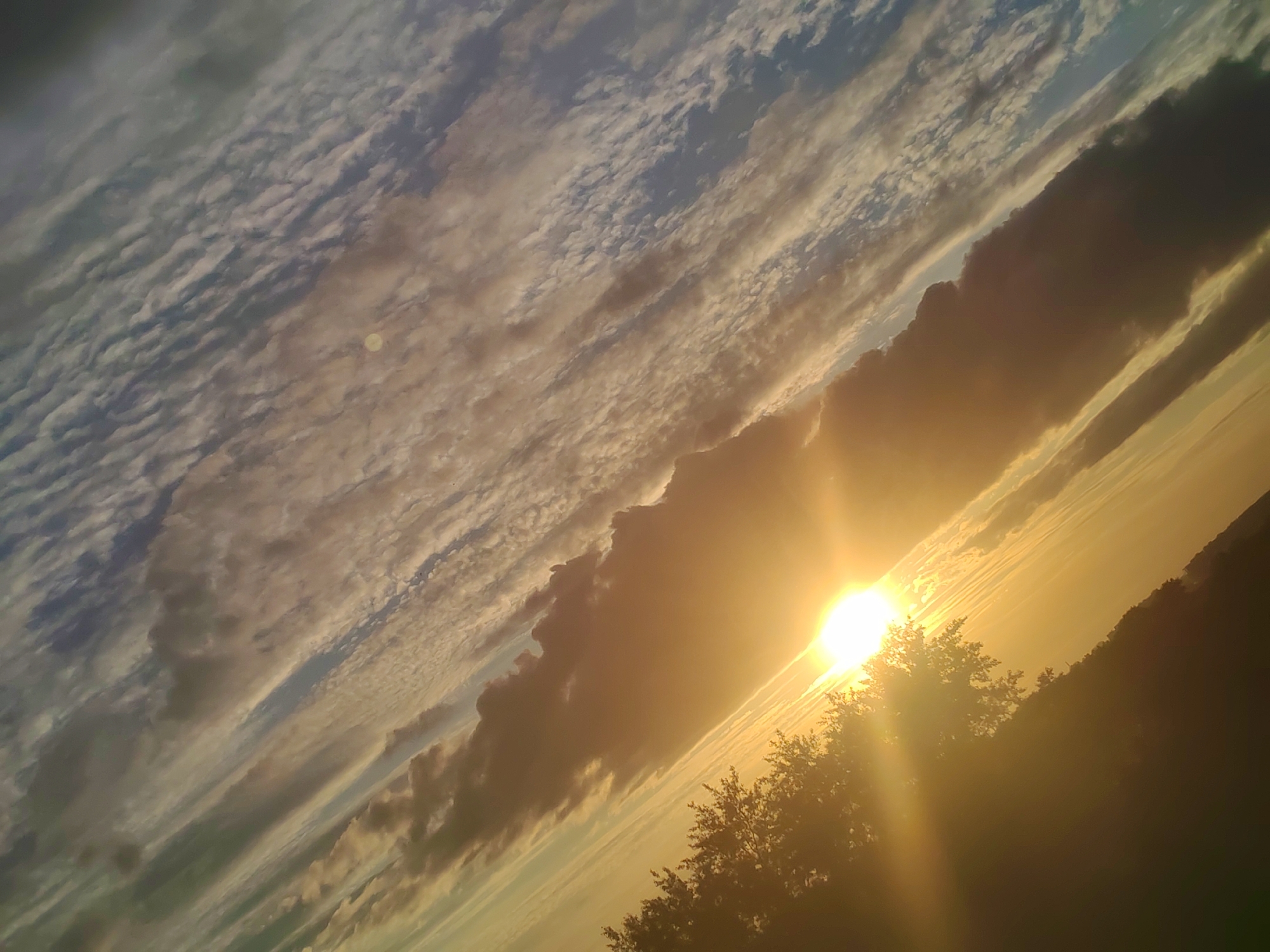 A diagonally oriented landscape image captures a vibrant sunrise or sunset over Podolsk, Russia, with the sun prominently featured. The brilliant sun is positioned in the lower right, emitting intense golden and orange light that creates a strong lens flare and bathes the scene in warm hues.
The sky is filled with a dynamic array of clouds. In the upper left, smaller, ripple-like cloud formations stretch across a lighter blue-grey portion of the sky. Towards the center and right, larger, darker cloud banks form dramatic silhouettes against the bright sunlight, some with illuminated edges and undersides. These clouds vary in density and form, creating a striking interplay of light and shadow across the expansive sky.
Along the lower right horizon, the dark silhouettes of trees are visible, standing out against the intense brightness of the sun and the glowing sky. The overall impression is one of natural beauty and peaceful grandeur, characteristic of either dawn or dusk. There are no visible human subjects, objects, or text in the image.