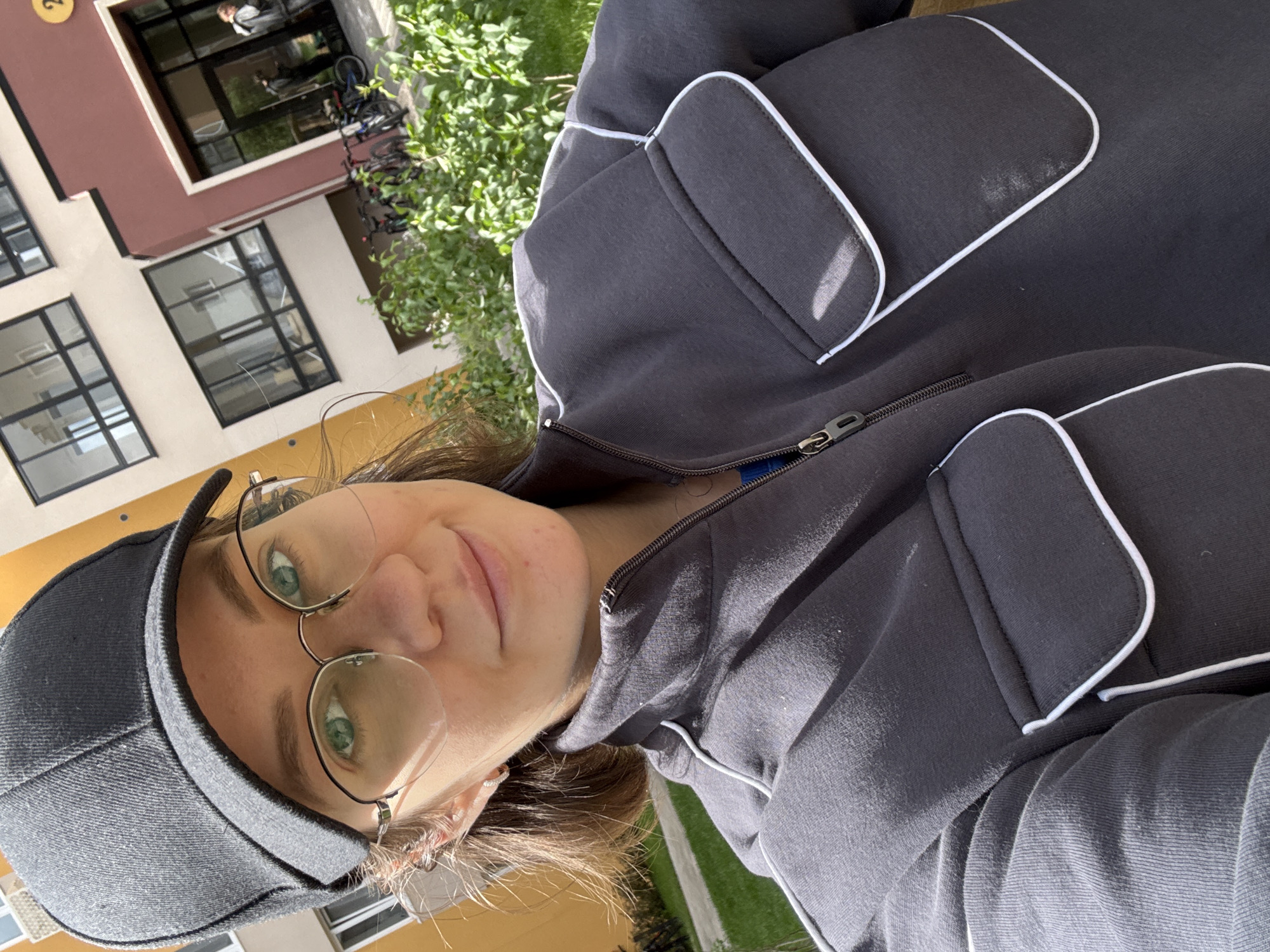A medium shot depicts a fair-skinned person with light brown hair wearing a grey baseball cap and wire-rimmed glasses, angled downwards toward the camera. The individual is wearing a grey zip-up jacket with white trim. The background features a multi-story building with beige and burgundy sections, including windows. A hint of green foliage and a glimpse of a bicycle stand are visible behind the building. The location appears to be outdoors during daylight. There is no conclusive evidence in the photo to identify the city or country. The city of Izhevsk, Russia, cannot be definitively determined as the location of this picture.