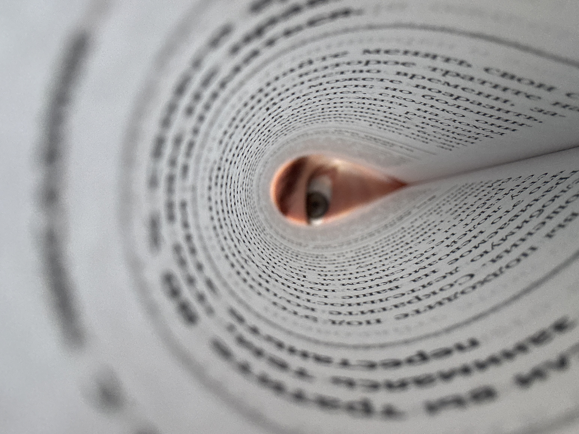 A close-up shot shows a book's pages rolled into a tube shape, creating a tunnel-like effect. Through the opening at the end of the tube, a person's eye is visible. The pages of the book are filled with text in an unknown language, and the image is well-lit. Location details are not discernible from the image.