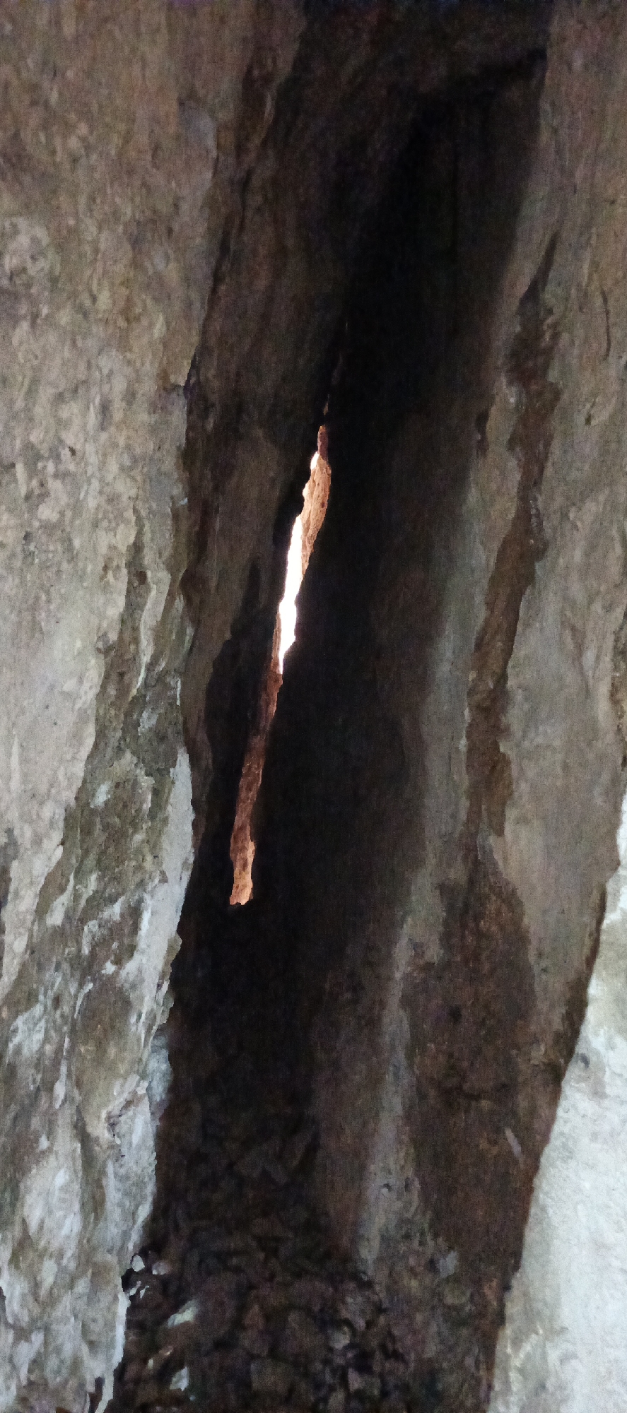 The image depicts a dark, narrow natural rock opening, possibly a cave entrance, a grotto, or a deep fissure in a rock face. No people or man-made objects are visible, making the primary subject the geological formation itself.
A significant detail is the bright, vertical, irregular slit within the darker opening, through which an intense light source is shining. This light casts a warm, reddish-orange glow on the edges of the crack, sharply contrasting with the deep shadows that dominate the rest of the cavity. This suggests it is daytime, with sunlight illuminating the passage from an external point.
The rock walls surrounding the opening are rough and uneven, displaying a variety of earthy tones including shades of grey, brown, and darker, almost black, areas in the deepest shadows. Lighter patches of tan or off-white rock are also visible where some ambient light might reach. The bottom of the opening appears to be composed of loose rocks or debris. The scene evokes a sense of natural exploration and the stark beauty of light penetrating darkness within a confined, rocky space. No visible text is present in the image.