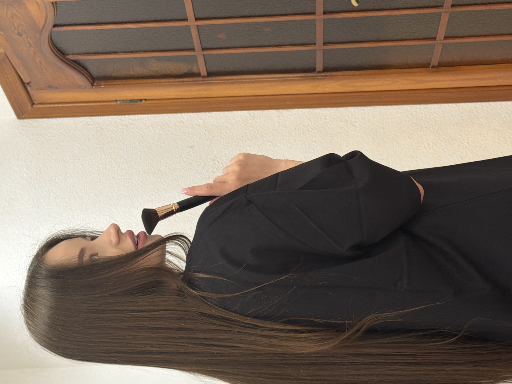 A person is shown from the side, holding a makeup brush to their lips. The person has long, brown hair and is wearing a black garment. The background consists of a white wall and a wooden window frame. The location cannot be determined to be in Kovel, Ukraine, based on the image alone.