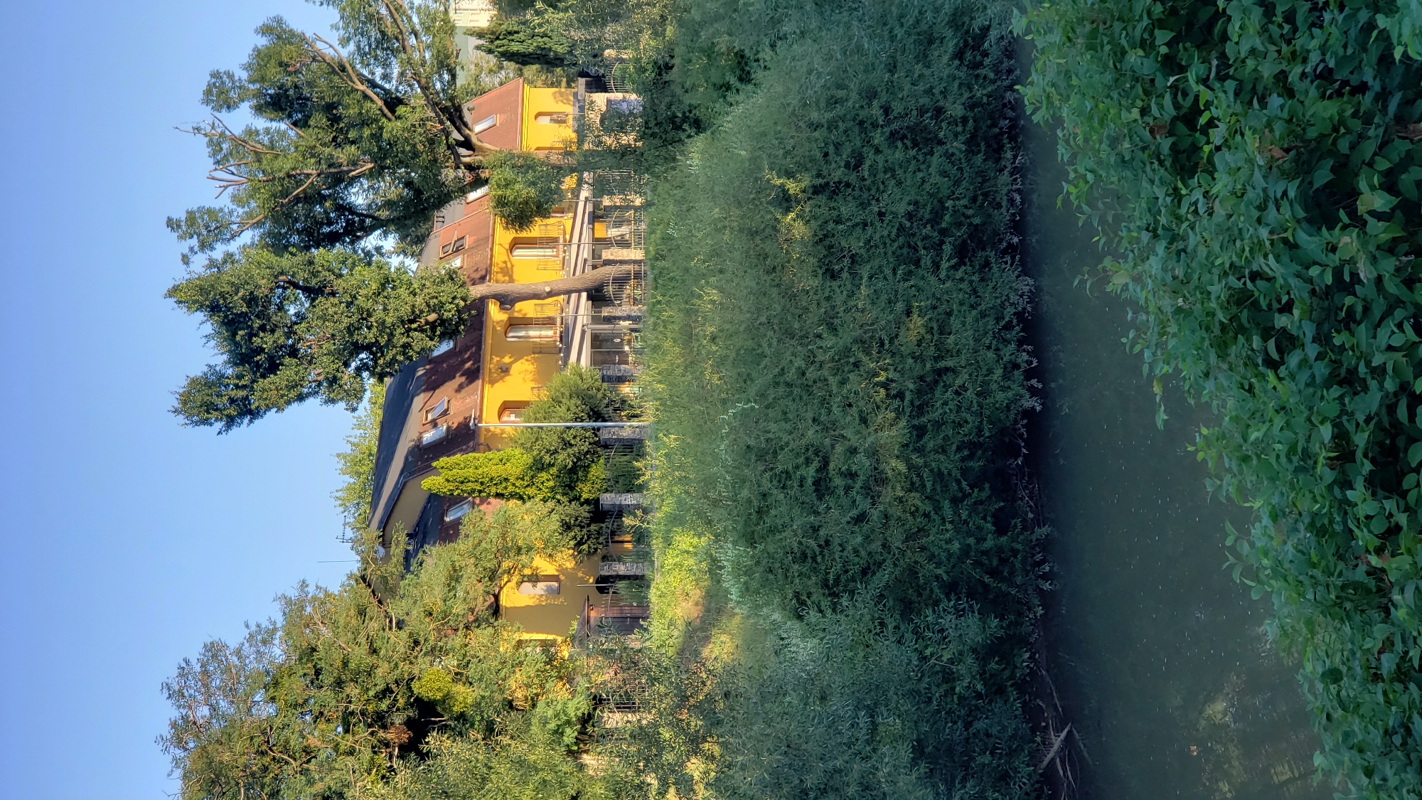 A sunlit, yellow-painted building with a red-tiled roof and dormer windows stands behind a line of lush green trees and bushes. The building appears to be a hotel or restaurant, with a glass-enclosed patio area visible. A dark, still body of water, likely a river, flows in the foreground, bordered by dense foliage. The scene is bathed in the warm light of what appears to be late afternoon or early morning. The location is near the Sekčov river in Prešov, Slovakia. There are no people visible in the image.