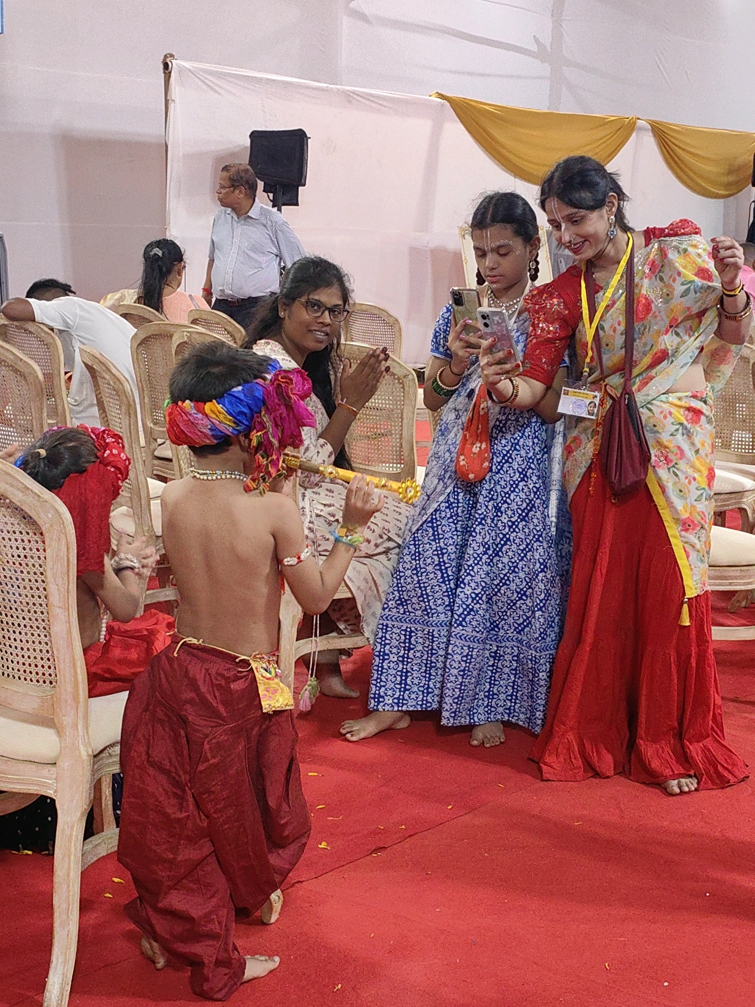 In Mumbai, India, at what appears to be a religious or cultural gathering, a group of people are observed in a brightly lit indoor space with a red carpeted floor. In the foreground, a young boy, possibly dressed as a deity, faces away from the camera, wearing a colorful turban and ornate red pants, playing a flute. To his right, two women stand looking at a mobile phone. One woman, in a red and floral patterned sari, is looking at the phone with a smile, while the other, in a blue and white patterned outfit, also appears engaged with the device. Further in the background, other individuals are visible, some seated, including a man in a striped shirt looking over his shoulder. The environment features simple white fabric backdrops and decorative yellow drapes. The overall atmosphere suggests a celebratory or communal event.