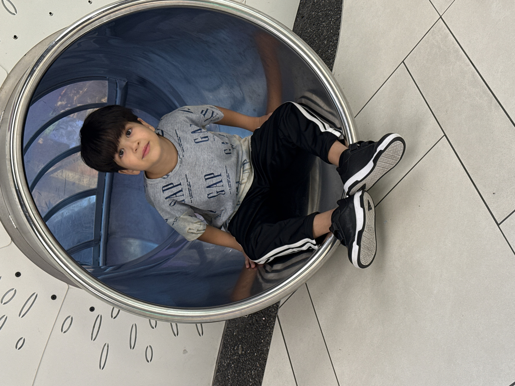 A young boy with dark hair and bangs sits inside a large, silver, cylindrical tube slide. He is wearing a gray t-shirt with the word "GAP" printed repeatedly in a darker gray font, and black athletic pants with white stripes down the sides. He has black sneakers on his feet. The boy is looking at the camera with a slight smile. The slide is situated on a light-colored tiled floor with dark grout lines. Behind the boy, the interior of the slide appears to be made of blue-tinted material. The exterior wall of the slide structure has a white surface with scattered oval and dot patterns. The background outside the slide is blurred and appears to be an indoor environment.