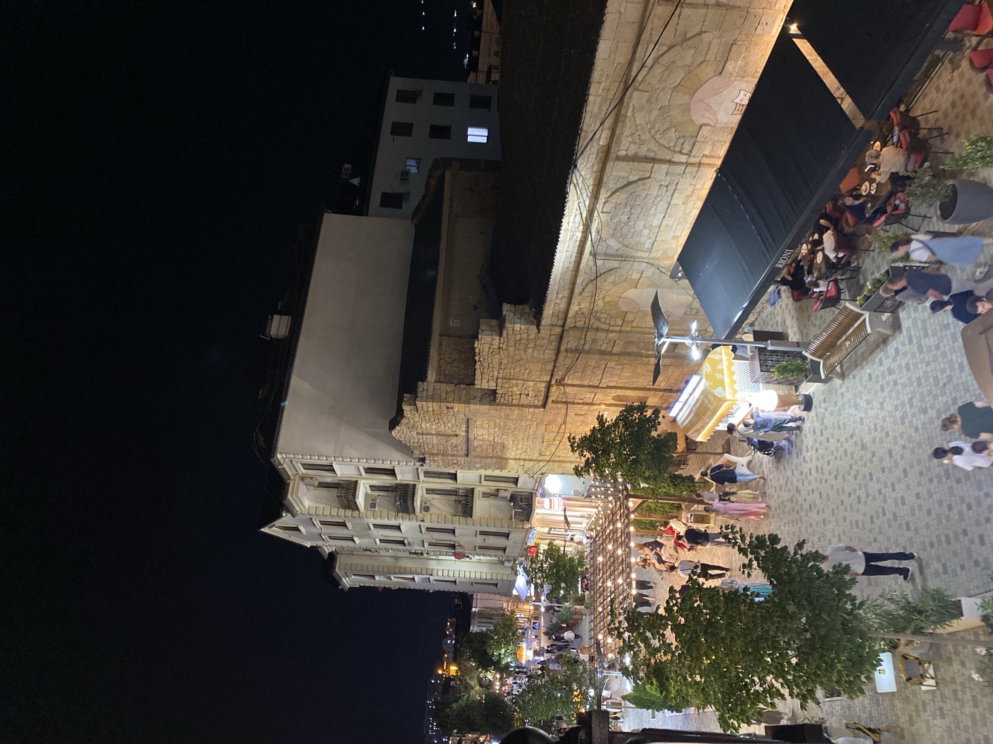 This image captures a nighttime scene on a bustling street in Derbent, Russia. The view is from an elevated perspective, looking down onto a pedestrian area lined with buildings and outdoor seating areas for establishments.
In the foreground, a section of a stone wall, likely part of an older structure, dominates the right side of the frame. Overhanging this wall is a black awning with the word "RIONI" visible in white lettering, suggesting a restaurant or cafe. Underneath this awning, tables are set with chairs, and several people are seated, appearing to be dining or socializing. Red chairs are visible at some of the tables.
Further into the scene, a paved pedestrian walkway meanders through the area. Several trees with green foliage are planted along the walkway, some illuminated by string lights draped over pergolas. People are walking along the path, some in groups, others individually.
On the left side of the image, a multi-story building with a light-colored facade and dark windows stands prominently. It has a distinctive pointed roofline and balconies. Other buildings are visible in the background, some with illuminated windows, suggesting residential or commercial use. The street is well-lit by a combination of streetlights and the lighting from the businesses.
In the middle ground, an ice cream stand with a yellow canopy is visible, with people queuing or being served. Various tables and chairs are also placed in open areas, inviting people to sit and enjoy the evening. The overall impression is of a lively and active urban environment, popular with locals and visitors alike. The lighting creates a warm and inviting atmosphere despite the darkness of the night.