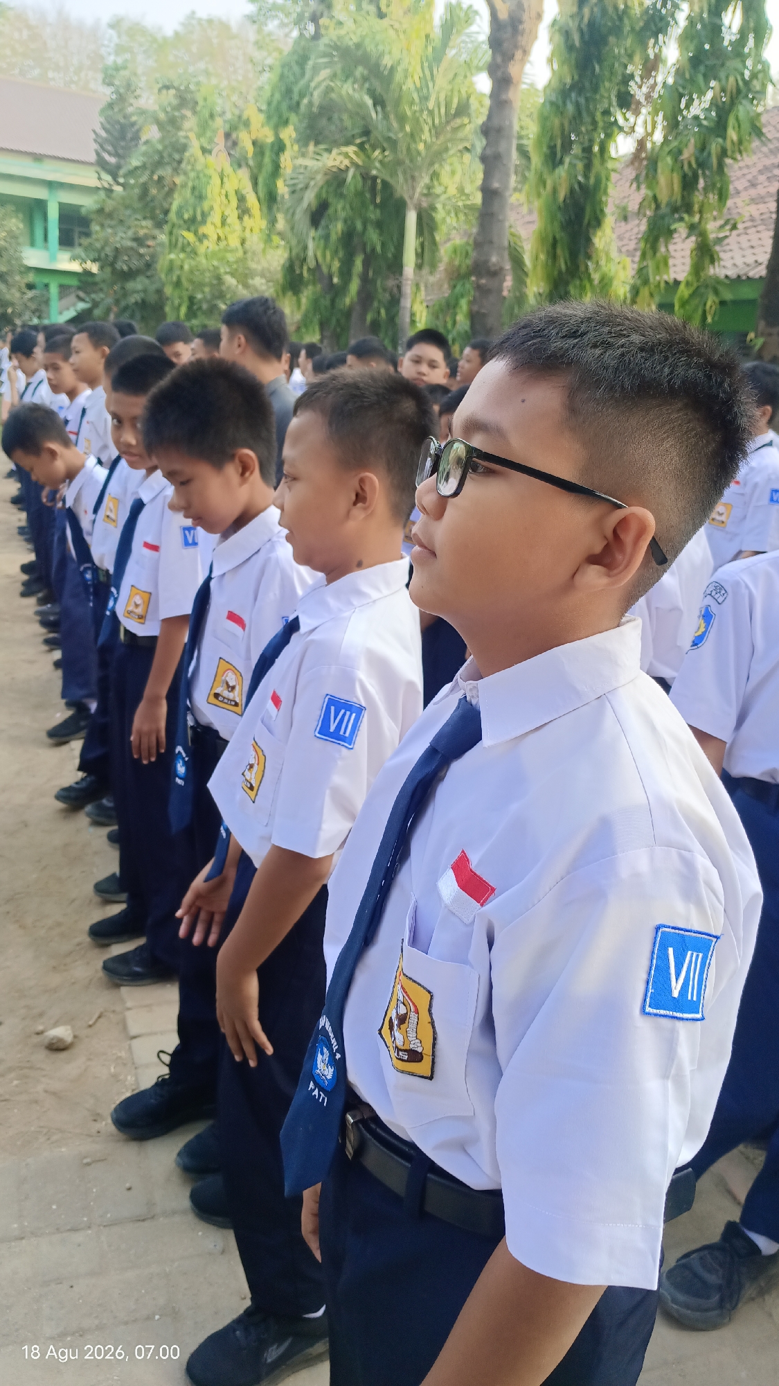 In Kecamatan Pati, Indonesia, a group of young male students in white shirts, blue ties, and dark pants stand in a line outdoors, facing away from the camera. They appear to be in uniform, indicated by matching patches on their shirts, including a red and white Indonesian flag emblem and a blue square with the Roman numeral "VII". One student in the foreground wears glasses and looks to the side. The background features lush green trees and a building, suggesting a school environment. The ground is dirt and paved. A timestamp in the lower left corner reads "18 Agu 2026, 07.00".
