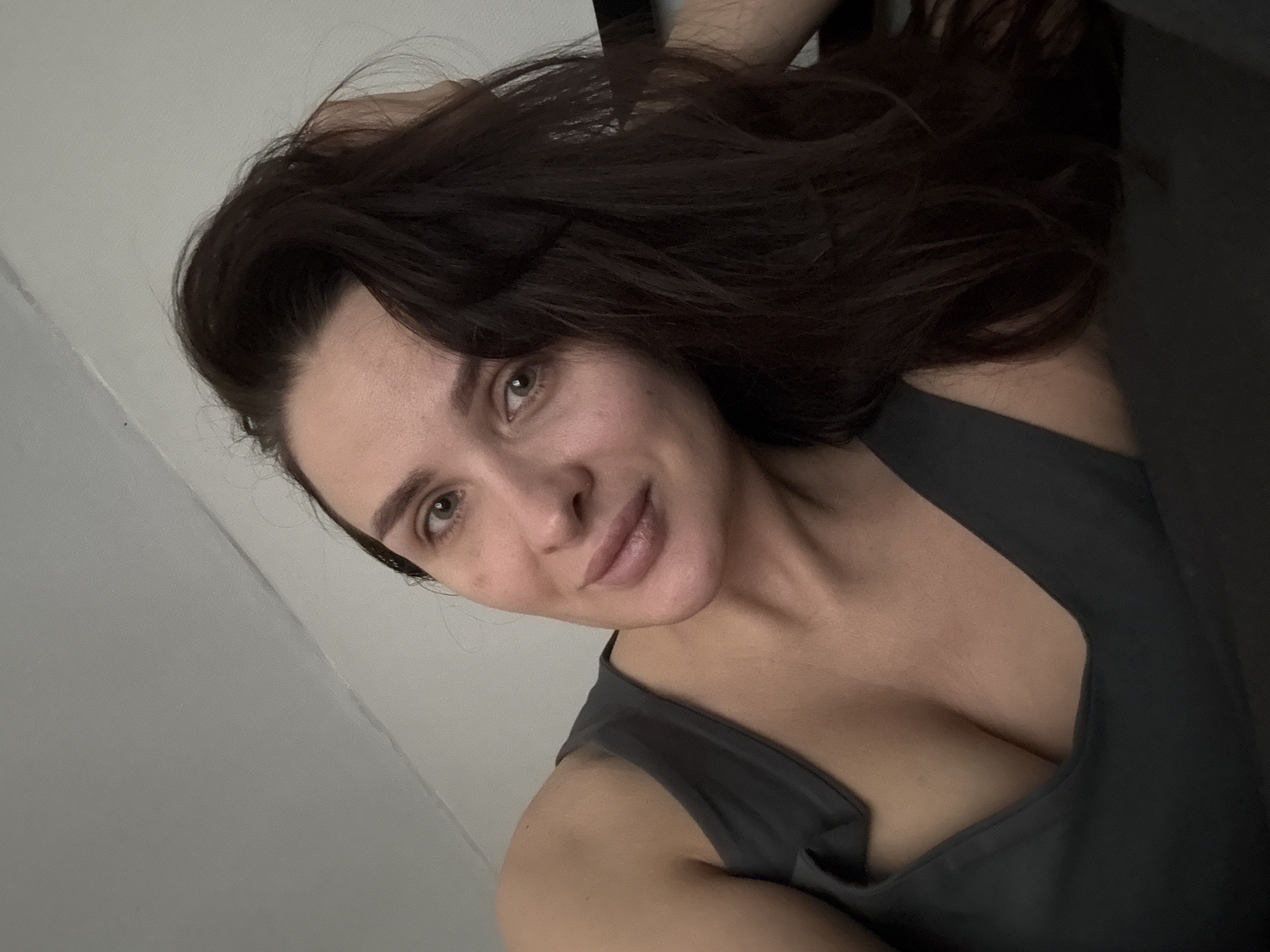 A woman with long, dark brown hair is posing for a selfie. She has fair skin, light brown eyes, and full lips. She's wearing a dark gray tank top with a deep V-neck. The background is a light gray wall. The location cannot be confirmed.