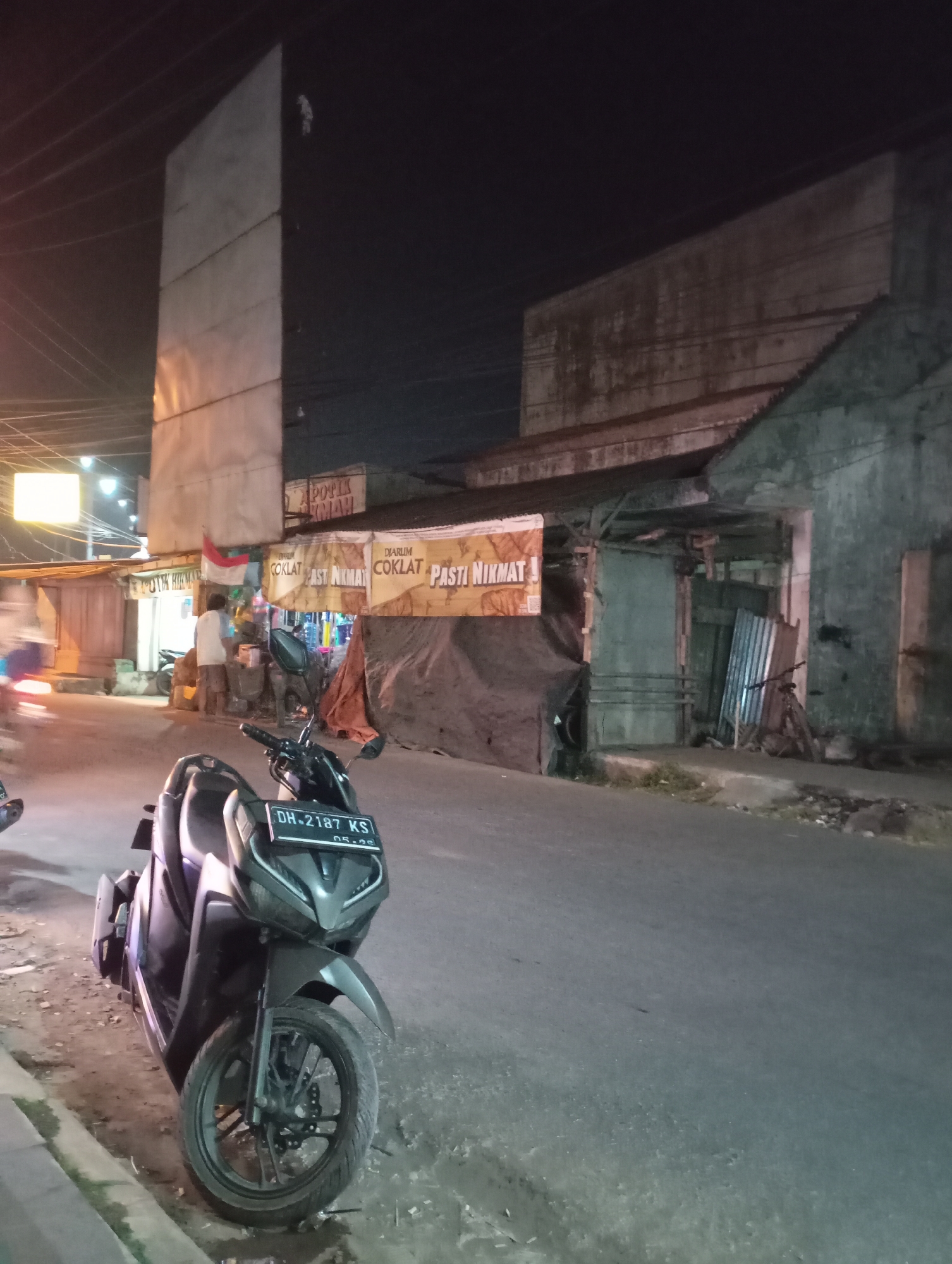 The image depicts a nighttime scene on a street in Kecamatan Haurgeulis, Indonesia. In the foreground, a dark-colored scooter is parked on the side of the road, facing left. The scooter has a license plate reading "DH-2187 KS".
Behind the scooter, the street continues, lined with several buildings. To the left, a tall, rectangular structure with a light-colored facade dominates the upper left portion of the image, with power lines running across the dark sky. Further down the street, a building with signage that appears to be in Indonesian is visible. The signage includes the words "DIARULM COKLAT PASTI NIKMAT" repeated on banners.
In front of one of the buildings, a person wearing a light-colored shirt and dark shorts is standing near a stall or shop. The area around the shops appears to be an open market or street-side vendor area. There are also tarps covering parts of the structures. The overall environment suggests a commercial area at night, with artificial lighting illuminating parts of the scene. The road surface is asphalt, and the ground beside the road is a mix of dirt, gravel, and some debris.