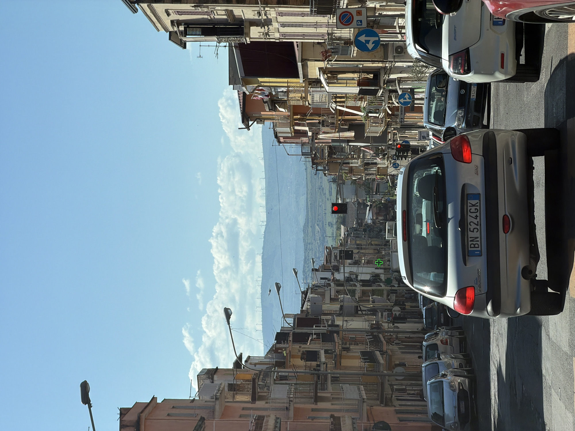 This vertical photograph captures a vibrant street scene under bright daylight in what appears to be an urban area, likely Scordia, Italy, as indicated by the context. The upper left of the image is dominated by a vast expanse of clear blue sky, adorned with scattered white clouds, suggesting fine weather conditions.
The lower right side of the frame features a bustling street with several vehicles. A silver Peugeot 206 hatchback, viewed from its rear, is prominent in the immediate foreground on the right. Its Italian license plate, "BN 524GK," is clearly legible. Behind it, other cars are lined up, including a white Suzuki Swift and a darker vehicle with a partial license plate, "CM 328YB." A red traffic light is visible on the right side of the road, midway into the scene. Further along, a green cross sign, commonly indicating a pharmacy in Italy, is also noticeable.
Multi-story buildings, characteristic of Mediterranean architecture with balconies, windows, and awnings, line what would be the right side of the street if the image were correctly oriented. Some laundry is visible hanging on a balcony. Streetlights extend from these structures over the roadway. Visible road signs include a blue circular sign with a white left-pointing arrow and a 'no parking' sign (a blue 'P' within a red circle and diagonal slash).
In the middle ground, beneath the expansive sky and nestled between the buildings, a hazy range of distant hills or mountains stretches across the horizon. Several wind turbines are visible along their crest. The scene is devoid of people, and the strong sunlight casts clear shadows, indicating a time of day with the sun relatively high in the sky.