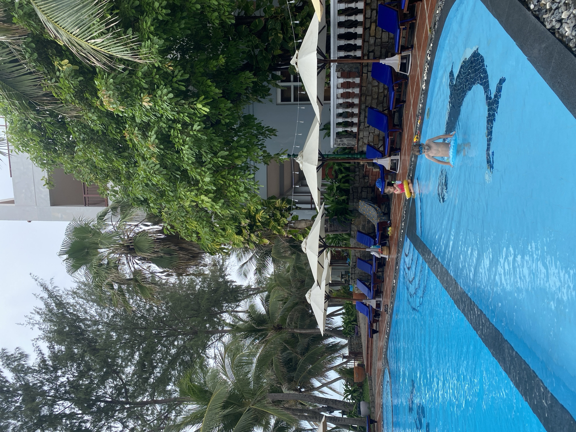 This image captures an outdoor swimming pool area, likely at a resort or hotel, during daytime under an overcast sky. The vibrant blue swimming pool dominates the right side of the frame, featuring a dark mosaic design resembling a dolphin or large fish on its bottom.
Two children are visible in the pool. An older child, wearing goggles and holding onto a blue inflatable ring, is partially submerged in the water near the center of the pool. Further to their left, a younger child is also in the water, wearing a pink and yellow inflatable ring. The children appear to be enjoying their time, playing or swimming.
Along the tiled edge of the pool, numerous blue lounge chairs are arranged, accompanied by white open umbrellas, suggesting a relaxation area. Beyond the pool and seating, lush tropical vegetation is abundant, including several tall palm trees with their distinctive fronds and dense green trees with rich foliage. In the background, white multi-story buildings, possibly part of the resort, are partially visible through the greenery. The general atmosphere suggests a leisurely vacation setting. No discernible text is visible in the image.