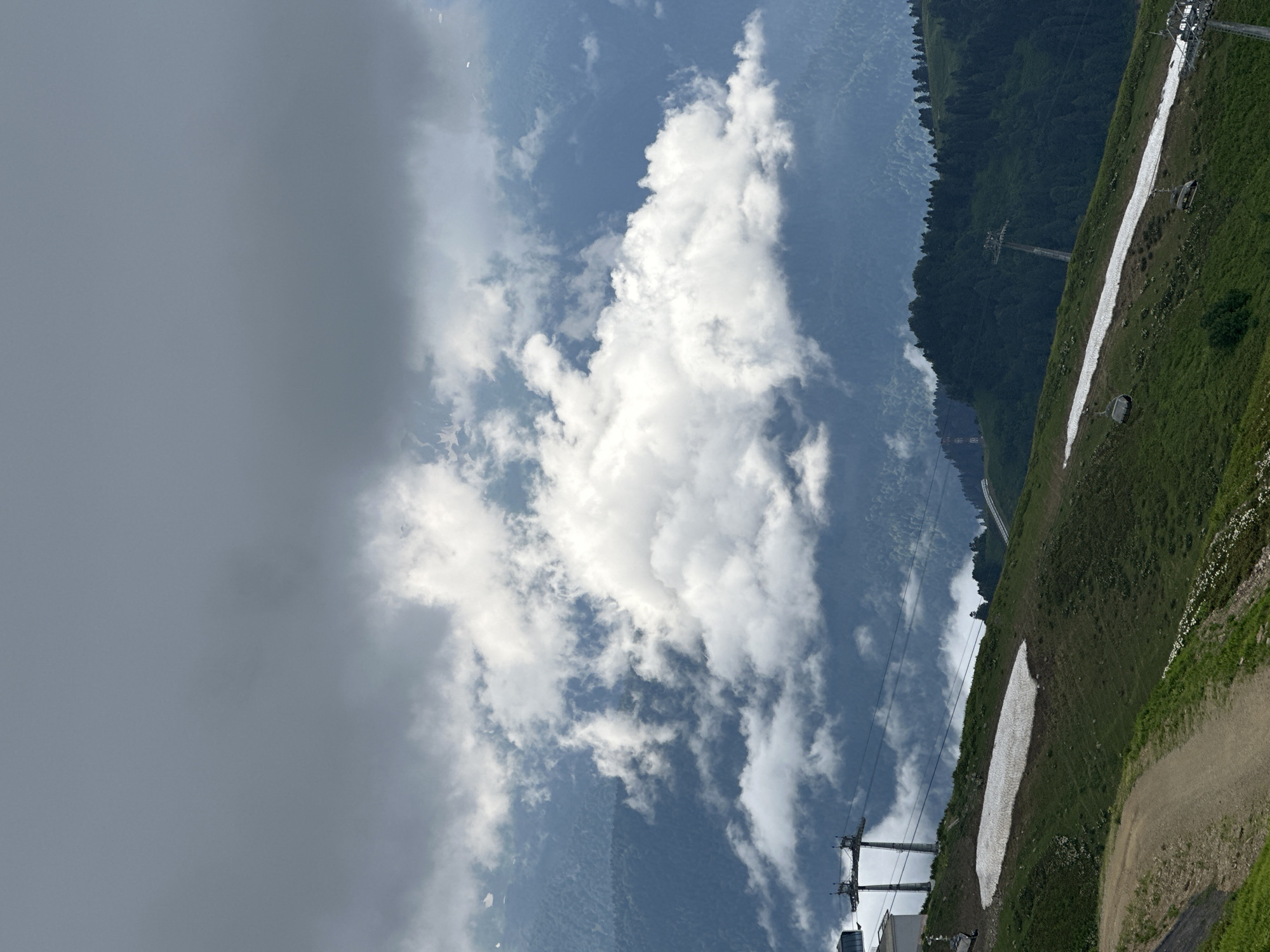 The image depicts a mountainous landscape in Estosadok, Russia, under a dramatic sky. In the foreground, a grassy slope features patches of snow and a dirt path winding down. Two ski lift towers are visible, one with a building attached to its base. The middle ground is dominated by lush green mountains covered in trees, with several more ski lift towers dotting the slopes. The background is a vast expanse of rolling mountains receding into the distance, partially obscured by a thick layer of white and gray clouds.