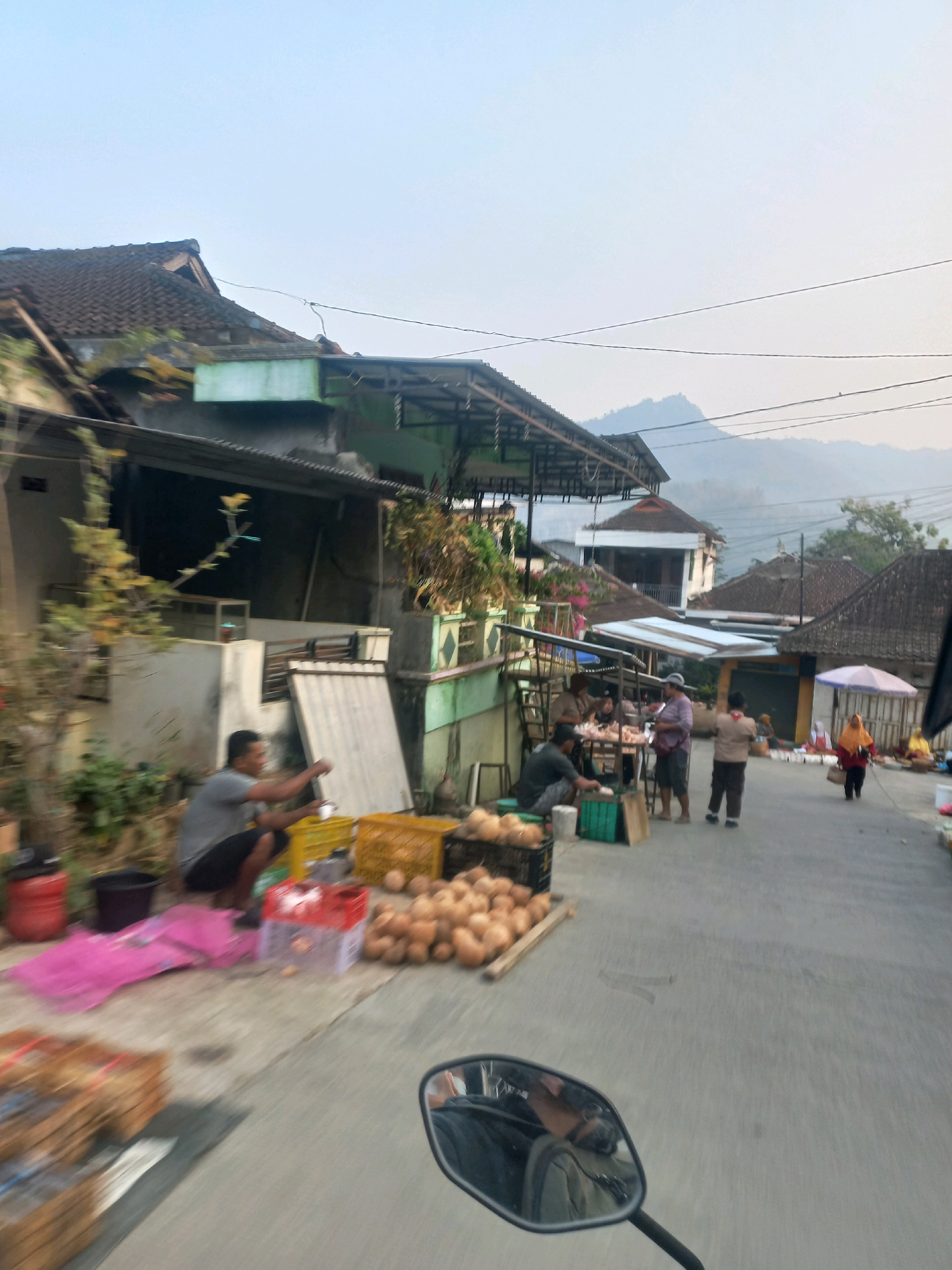This image captures a street scene in Sukoharjo, Indonesia, possibly a market. In the foreground, a motorcycle's side mirror is visible, reflecting a person wearing a grey shirt and dark shorts, crouching down. To the left, there are several stacked wooden crates, a red bucket, and a pile of pink fabric. Directly behind the crouching figure, a large quantity of coconuts are displayed in black crates and on the ground, some are on wooden planks.
Further down the street, there are several people engaged in activities related to a market. On the right side, a person wearing a yellow hijab and orange top walks away from the camera, carrying a white bag. An umbrella provides shade. Further back, a group of people are gathered around tables and stalls, possibly selling goods. Buildings with tiled roofs line the street, some appearing to be shops or houses. In the background, hazy hills are visible under a light sky. The overall atmosphere suggests a typical day in a local Indonesian town or city.