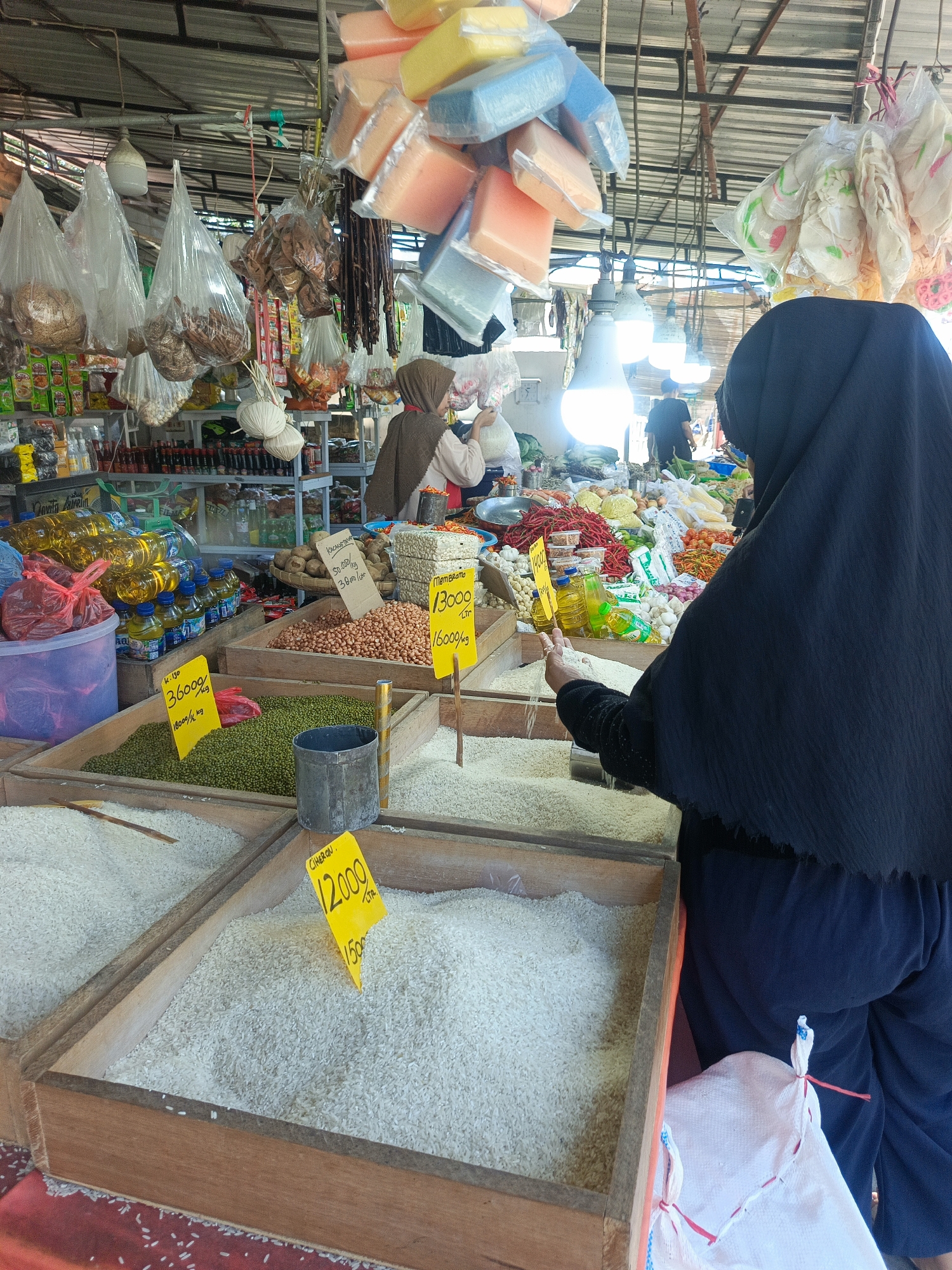 This image depicts a market stall in Tenggela, Indonesia, likely selling various food staples and dry goods. In the foreground, a woman wearing a black hijab and dark blue pants is shown from the back. She is standing in front of several wooden bins filled with rice. Her right hand is reaching into one of the bins, sifting through the rice.
The bins are labeled with yellow signs indicating prices. One sign reads "12000 Ltr," and below it "1500". Another sign, partially obscured, shows "36000 18000/kg". Behind the rice bins, there are containers of dried goods, including what appears to be lentils or small beans, and a basket of potatoes. Further back, bottles of cooking oil are stacked. The market stall is covered by a corrugated metal roof, supported by beams. Various items are hanging from the ceiling, including what looks like dried sausages, spices, and packaged snacks. In the background, other market-goers and stalls are visible, along with overhead lighting. The overall impression is a busy, traditional market environment.