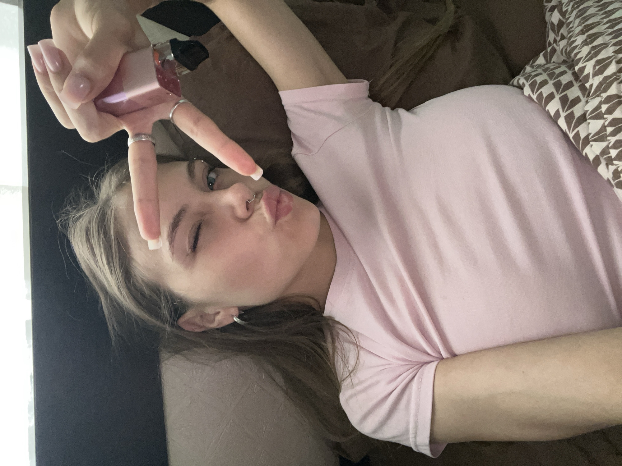 A young, fair-skinned woman with light brown hair, wearing a pink t-shirt, is lying on a brown surface. She is making a peace sign with her left hand while holding a small, possibly cosmetics-related, rectangular container between her fingers. She has multiple rings on her fingers and a piercing in her nostril. Her eyes are blue and she is winking with one eye and pursing her lips. A patterned cushion or pillow is visible on the right side of the image. A dark surface, possibly a wall, is visible on the left side. It is impossible to determine the location in the city of Фоки, Russia.