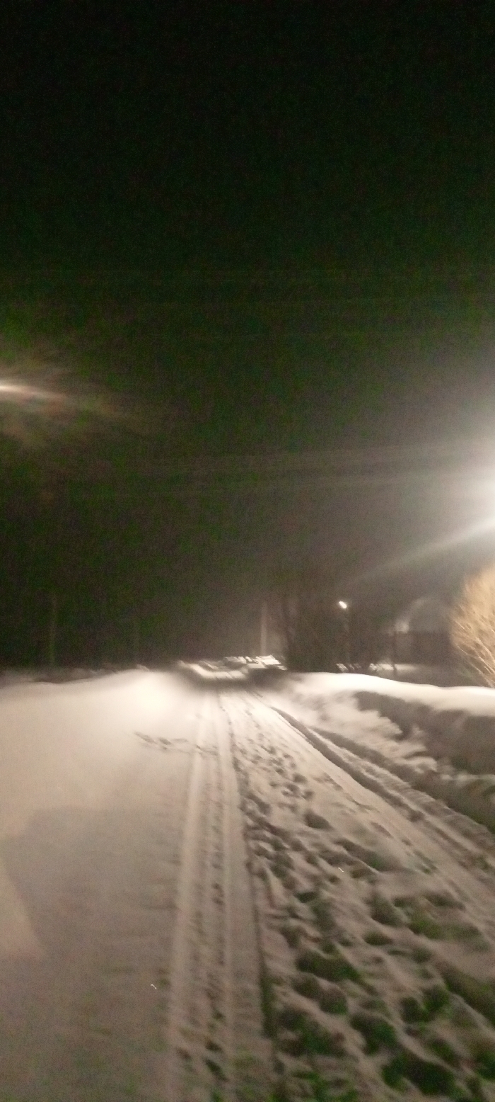 The image captures a snow-covered street at night in Красный Маяк, Russia. The environment is dark, illuminated primarily by artificial light sources. A prominent streetlamp on the right casts a strong, hazy light, creating significant lens flare in the upper right portion of the frame. Another diffused light source is visible on the far left. The foreground features a wide snow-covered path or road with distinct tire tracks running along the center-left and numerous footprints embedded in the snow, particularly on the right side of the tracks. Snowdrifts or banks line the edges of the path. In the midground and background, blurred outlines of bare trees and low-lying structures or buildings are discernible against the dark sky, suggesting a rural or suburban setting. No individuals or active subjects are visible in the scene.
