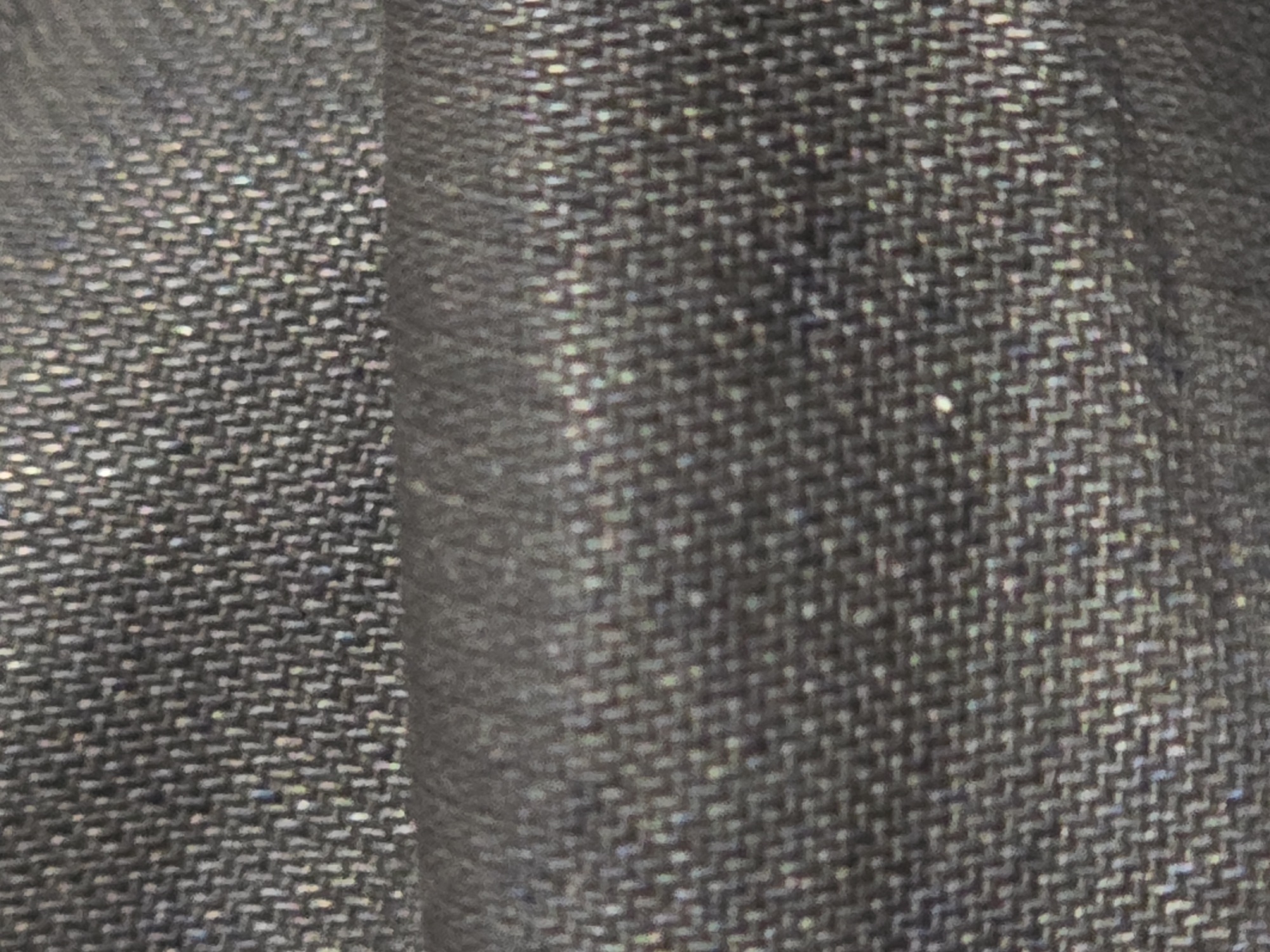 The image is a close-up, abstract photograph of a textured, dark gray fabric. The fabric appears to have a woven pattern, possibly a herringbone weave, with subtle iridescent flecks that catch the light. The lighting is diffused, creating a soft shadow in the lower half of the image where the fabric folds. There are no people, discernible objects, or environmental context visible. The image is an extreme close-up of a textile.