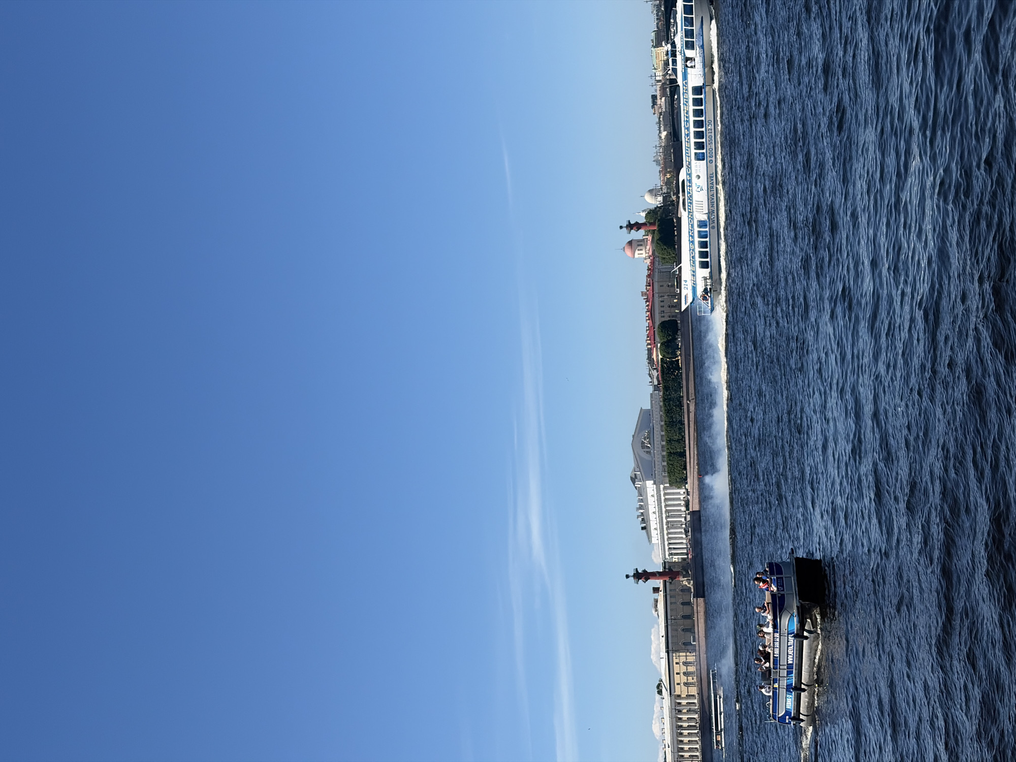 The image depicts a daytime scene on the Neva River in St. Petersburg, Russia, looking towards the Strelka of Vasilyevsky Island. The upper two-thirds of the image show a clear, bright blue sky with a few faint, horizontal wispy white clouds. The lower third consists of dark blue, choppy water, exhibiting white foamy wakes from moving vessels.
In the mid-ground, two boats are visible on the water. To the lower left, a smaller, flat-bottomed boat, colored dark blue and grey, moves from left to right. Approximately 6-8 individuals are seated in this boat, facing forward or slightly to the right. Text including "NEWHK-1" and "WWW.NEVA.TRANS" is visible on its side. To the lower right, a larger, white and blue high-speed hydrofoil-type boat, numbered "214," travels from right to left, generating a significant white wake. The side of this boat displays Cyrillic text "ПЕТЕРГОФ • КРОНШТАДТ • ОРЕШЕК • СТРЕЛЬНА" and "WWW.NEVA.TRAVEL." One person is partially visible near the front cabin.
The background features the Strelka of Vasilyevsky Island. Prominently centered are two tall, reddish-orange Rostral Columns. Between them stands a large, white neoclassical building with a colonnade, identified as the Old Stock Exchange. To the left of the left Rostral Column, a series of long, multi-story buildings with yellowish-tan facades line the embankment. To the right of the right Rostral Column, other buildings are visible, including one with a distinct reddish-pink dome and another with a smaller white dome. Sparse patches of dark green foliage are present along the riverbank. Faint structural elements of a distant bridge are discernible in the far right background.