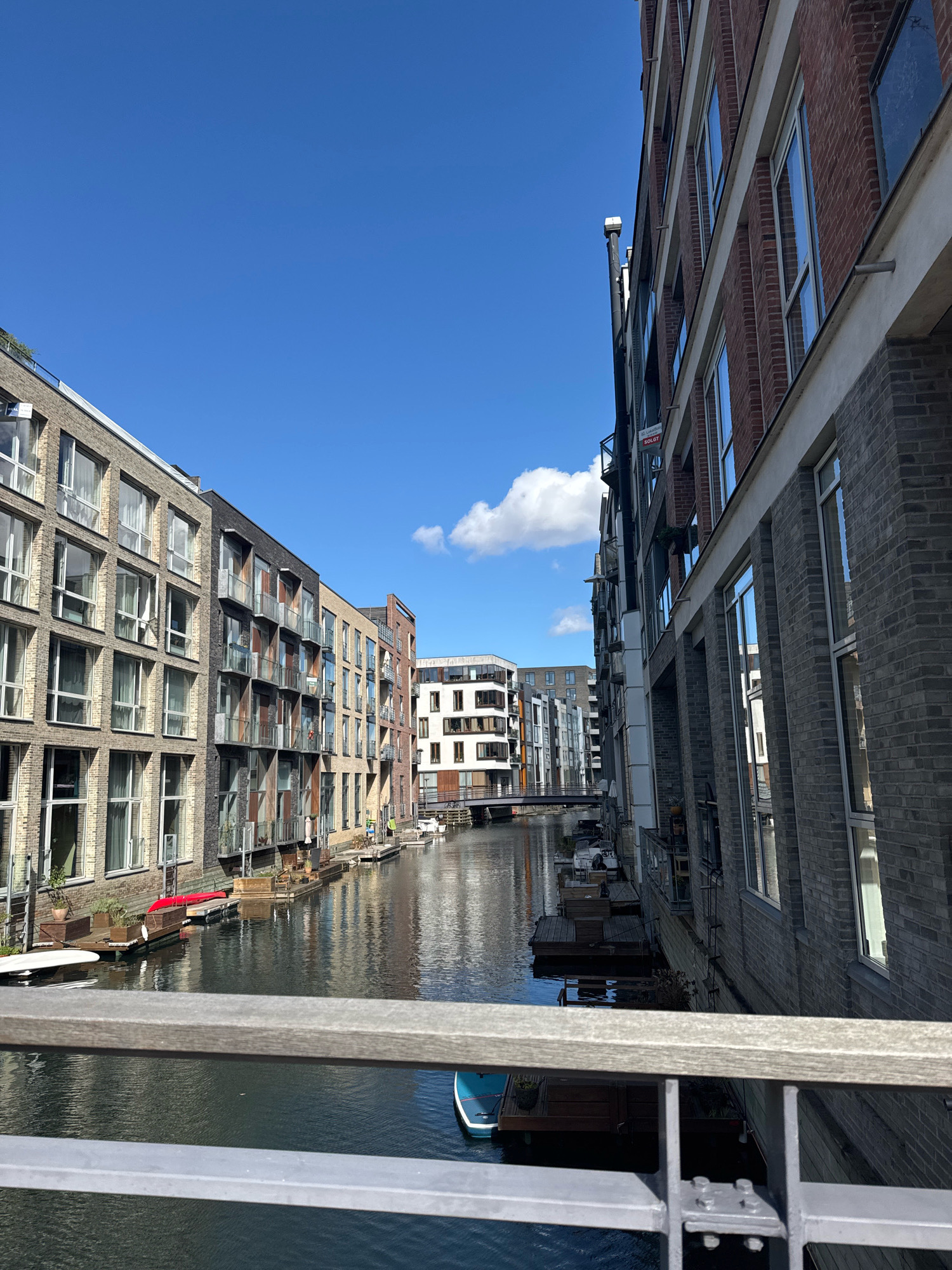 A bright, sunny day illuminates a canal lined with modern brick buildings. The canal is calm, reflecting the blue sky and the buildings. On the left, a row of light-colored brick buildings with many windows and balconies face the water. To the right, a darker brick building with large windows is prominent. Several small boats, including a red canoe-like vessel and a blue paddleboard, are docked along the canal's edge. A pedestrian bridge can be seen in the distance. A metal railing runs across the foreground, obscuring the lower portion of the scene. The sky is a clear, vibrant blue with a few small white clouds.
