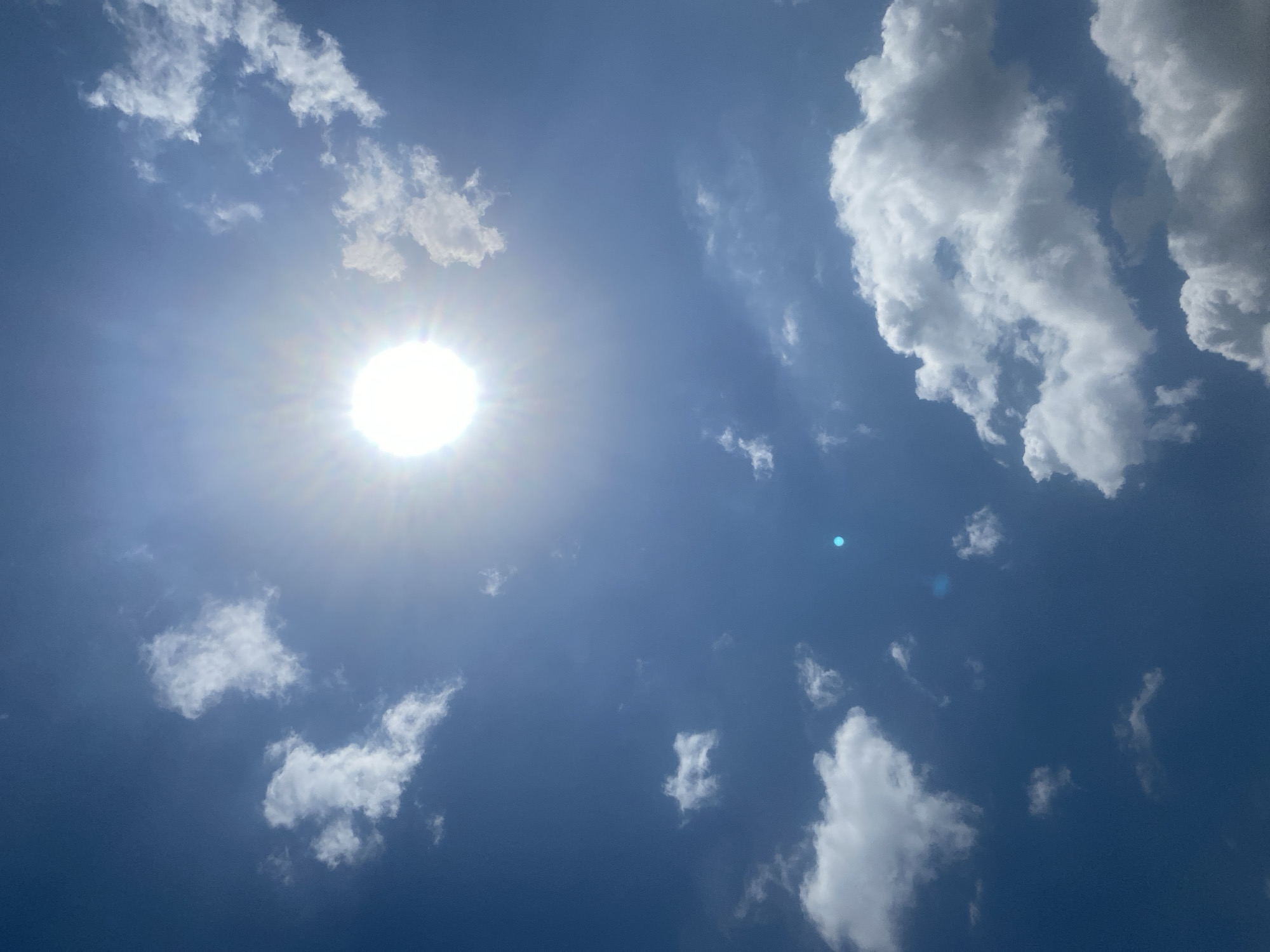 The image depicts a bright, sunny sky with scattered white clouds. The sun is prominently visible in the upper left quadrant of the image, emitting a strong lens flare. The sky is a deep blue, suggesting a clear day. The clouds vary in size and density, with some appearing wispy and others more voluminous. One distinct, larger cloud formation is visible in the upper right corner, with sharp edges and shaded undersides, indicative of its thickness and the angle of the sun. In the center of the image, a smaller, faint blueish dot is present, possibly a reflection or artifact. The overall scene conveys a sense of a bright and clear daytime atmosphere, typical of a warm climate. There is no visible text, people, or specific objects other than clouds and the sun. The setting appears to be an outdoor environment under a vast sky, consistent with the provided location context of Lafia, Nigeria. The lighting and the presence of sun and clouds suggest it is daytime, likely mid-morning to afternoon.