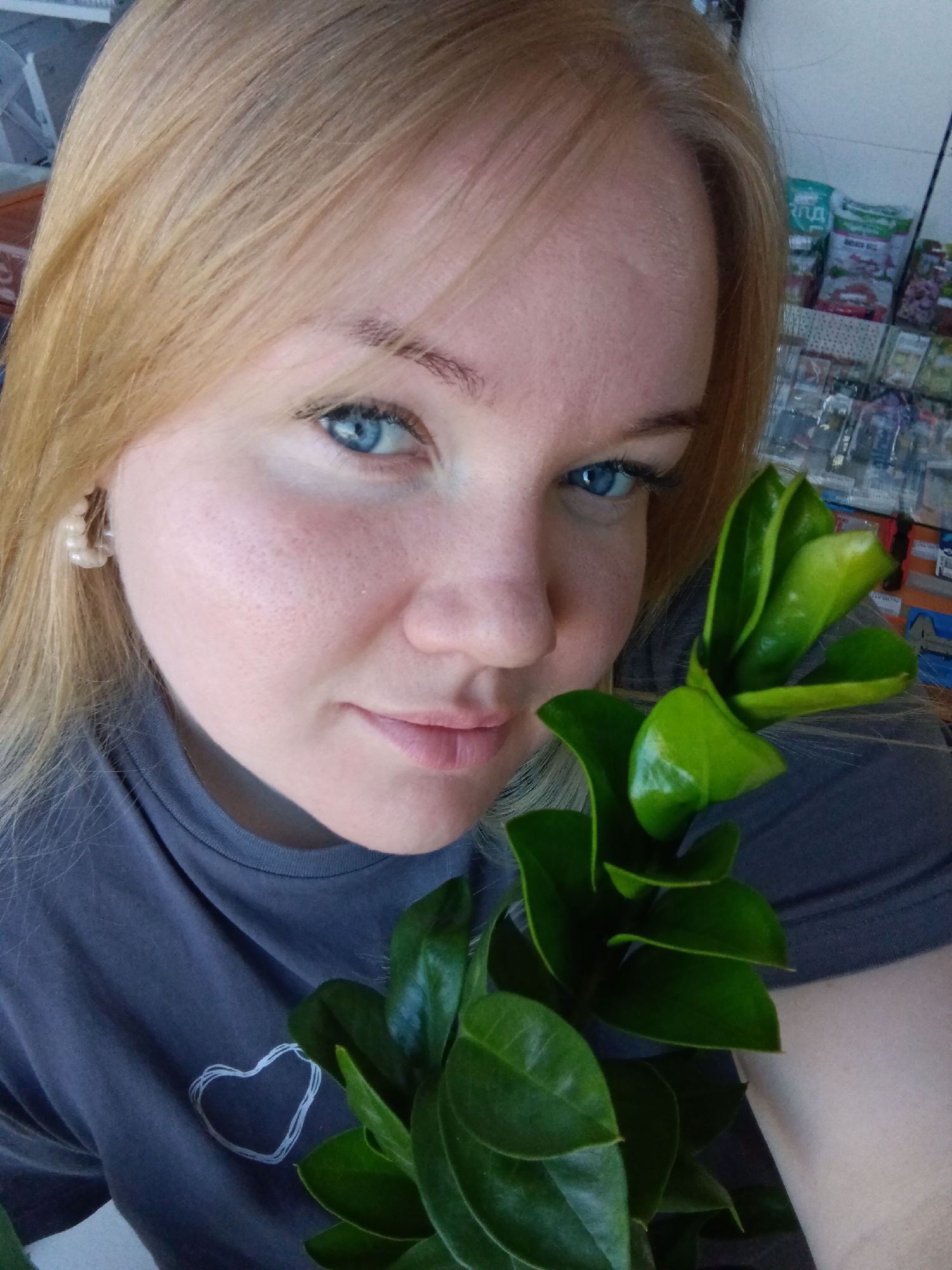 A close-up shot depicts a woman with blonde hair and blue eyes. She is wearing a dark grey t-shirt with a white heart outline design. She is holding a branch with green leaves. Behind her is a display of what appears to be seed packets. The location is identified as Фоки, Russia. The environment suggests an indoor setting, possibly a store or a home setting.