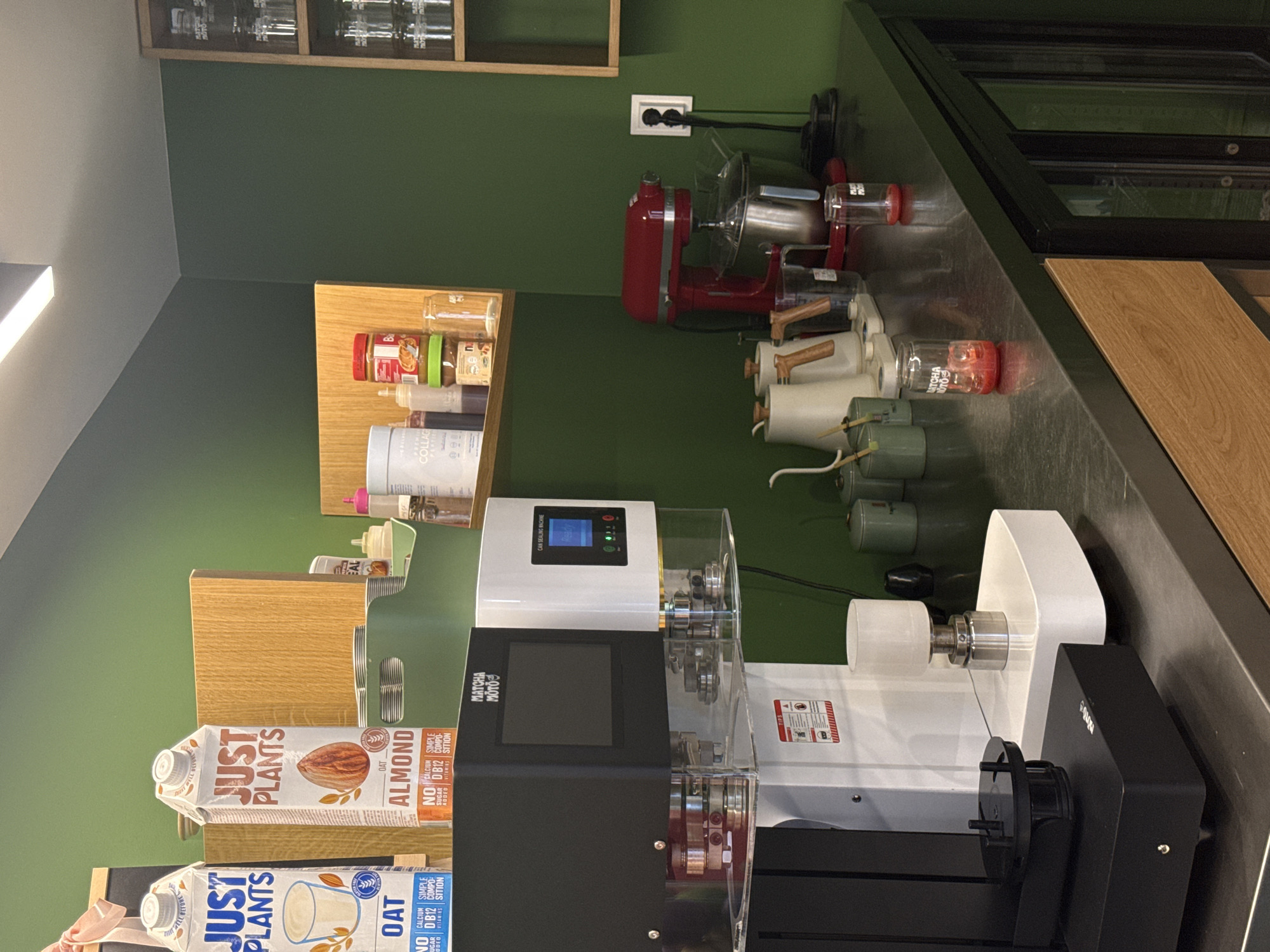 The image shows a section of a commercial kitchen with a focus on food preparation equipment and storage. There are several pieces of equipment on a dark countertop. These include a red stand mixer, multiple electric motors (possibly for food processing), and two can sealing machines, one white and one black.
The backdrop is a green wall with wooden shelving. On the shelving are various food items including nut butters, sauces, and containers. Below the lower shelf, there are two cartons of "Just Plants" brand milk alternatives; one almond milk and one oat milk.
The environment suggests a professional food-related establishment. There is no specific indication of Sofia, Bulgaria.