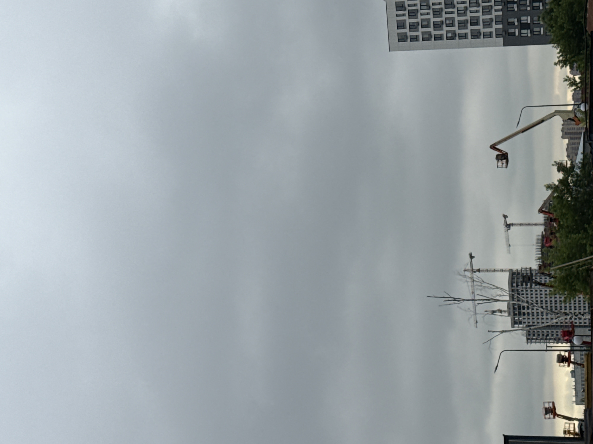 The image depicts an overcast sky above a construction site in Tomilino, Russia. In the foreground, a modern, multi-story apartment building is visible on the right, featuring a facade of numerous windows and sections of gray and white cladding. To the left of the apartment building, a boom lift with a basket is positioned, its arm extended. Further left, another boom lift is partially visible, along with parts of buildings under construction, including cranes and the skeletal framework of a high-rise building adorned with a grid of windows. Several trees and greenery are present in the lower section of the image, with a lamppost also visible. The overall scene suggests ongoing urban development under a cloudy sky.