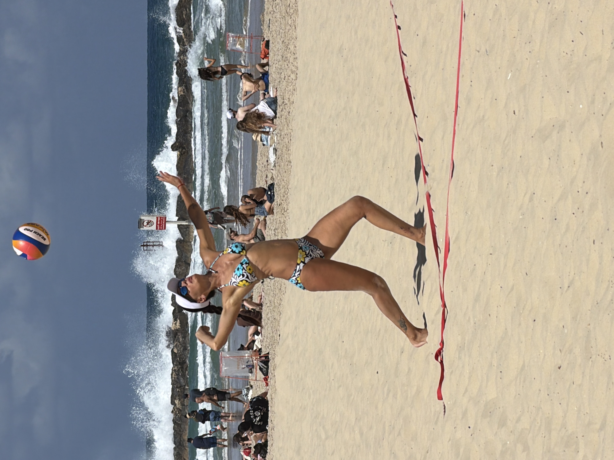 A woman in a floral bikini and a white cap plays beach volleyball on a sunny day in Tel-Aviv, Israel. She is about to hit a multi-colored volleyball. A red net is to her right. In the background, behind the volleyball court, the Mediterranean Sea crashes against a rocky breakwater. People are visible on the beach, some walking near the water, others sitting and watching the volleyball game.