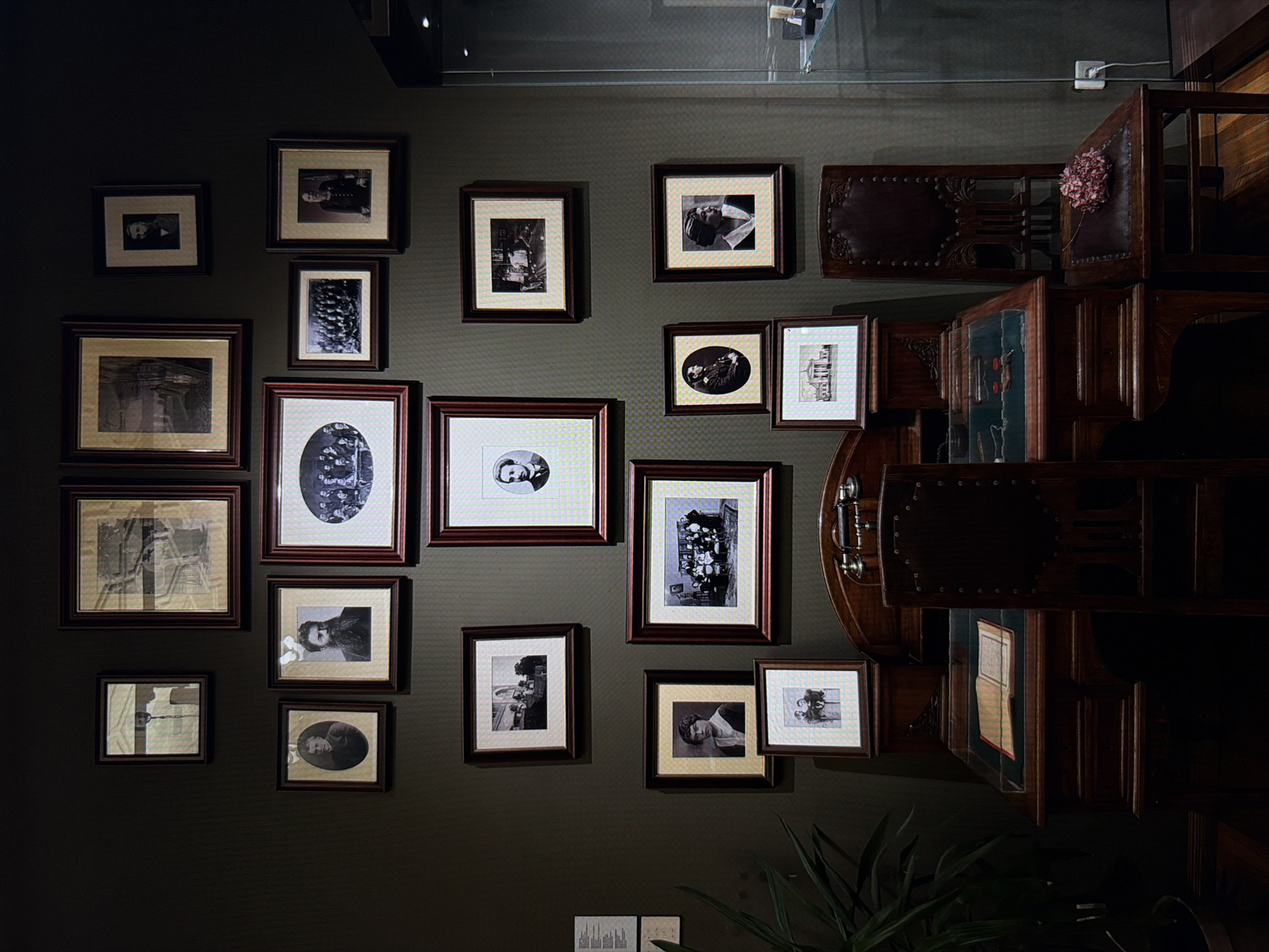 The image depicts a collection of framed black and white photographs displayed on a dark, textured wall. The photographs appear to be vintage, featuring portraits of individuals, groups, and some architectural scenes. The frames are predominantly dark wood, with some having a lighter matting.
The arrangement of the photographs is varied, with some hung individually and others in clusters. In the upper right portion of the image, part of a dark wood cabinet or display case is visible, adorned with ornate metal detailing and what appears to be a dried flower. This cabinet has glass panes, revealing some objects within, possibly books or decorative items. The overall lighting suggests an indoor setting, likely dimly lit with focused illumination on the displayed items. The mood is subdued and possibly nostalgic, given the nature of the photographs. There is no visible text within the photographs or on the wall. The scene gives the impression of a curated display, perhaps in a museum, gallery, or a personal collection within a home. The photographs are primarily portraits of people in formal attire, hinting at a historical context. Some images contain multiple individuals, suggesting family gatherings or formal sittings. The presence of a plant in the lower right corner adds a touch of life to the otherwise static display.
