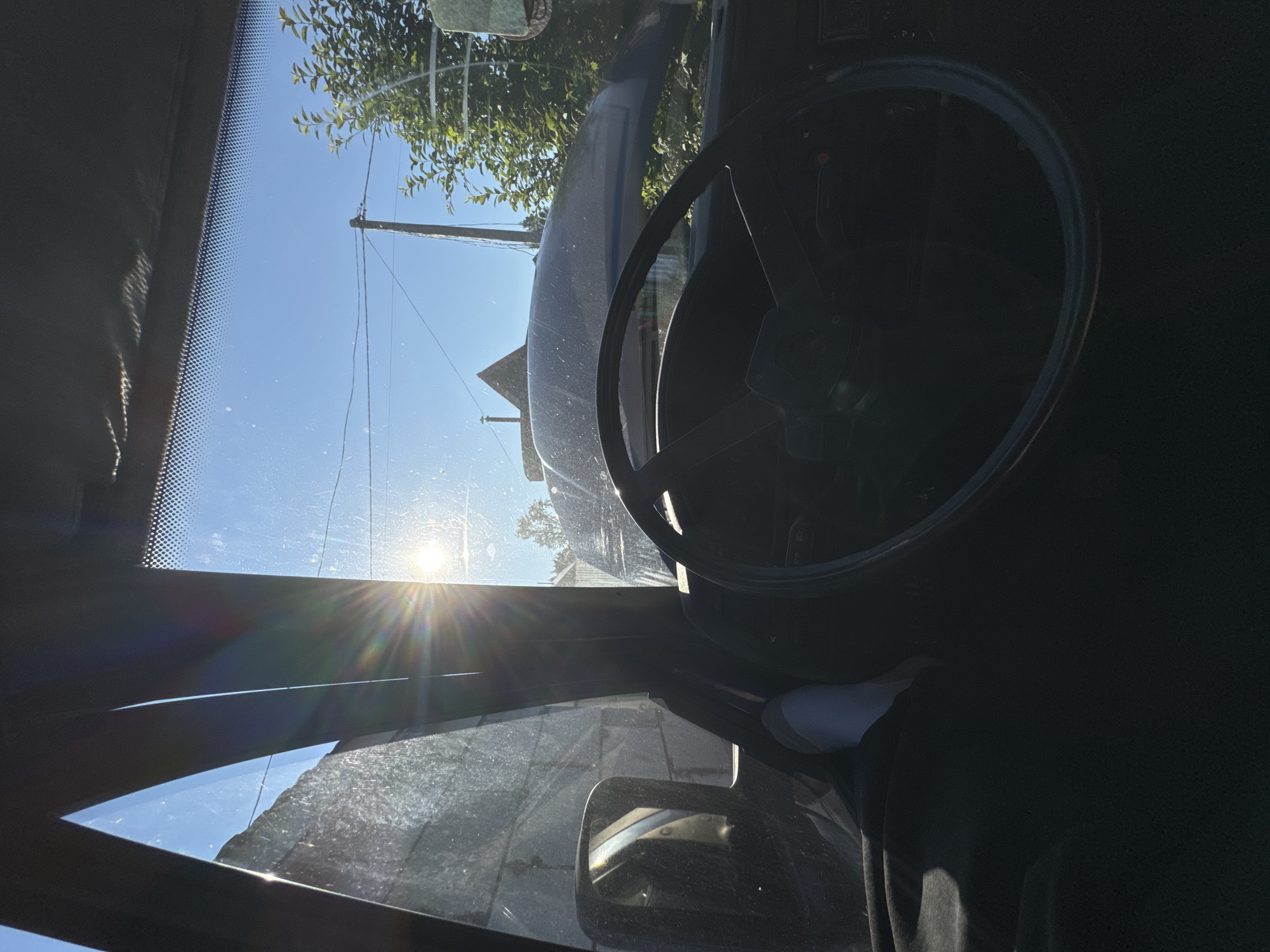 The image, taken from inside a vehicle, shows a sunlit exterior with a clear blue sky. The sun is prominent, creating a strong lens flare. The view through the windshield and side windows reveals a somewhat dusty environment with a rough-textured wall on the left and trees and a utility pole in the distance. The interior of the vehicle is dark, with a steering wheel visible in the foreground. A partial view of a person's foot, wearing a white sock, is also visible inside the car. The overall impression is of a still, sunny day, possibly in an outdoor or semi-rural setting within Krasnokamsk, Russia.
