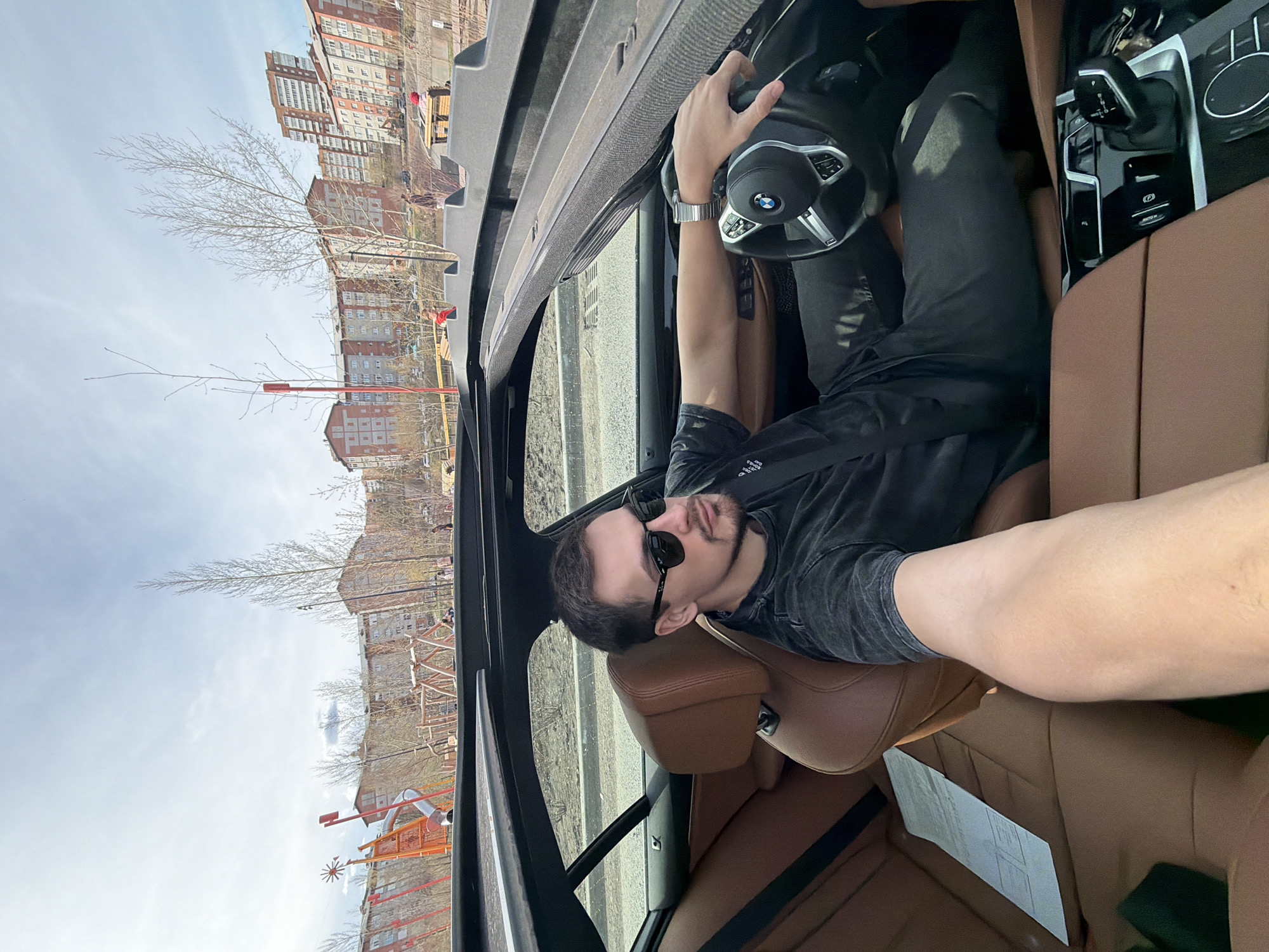 A man sits in the driver's seat of a car, wearing a dark-colored shirt, sunglasses, and a wristwatch on his left wrist. He holds the steering wheel, and his seatbelt is fastened. The car has brown leather seats. Outside the car window, there is an urban landscape with multi-story brick buildings and a playground. The setting is Krasnoyarsk, Russia.