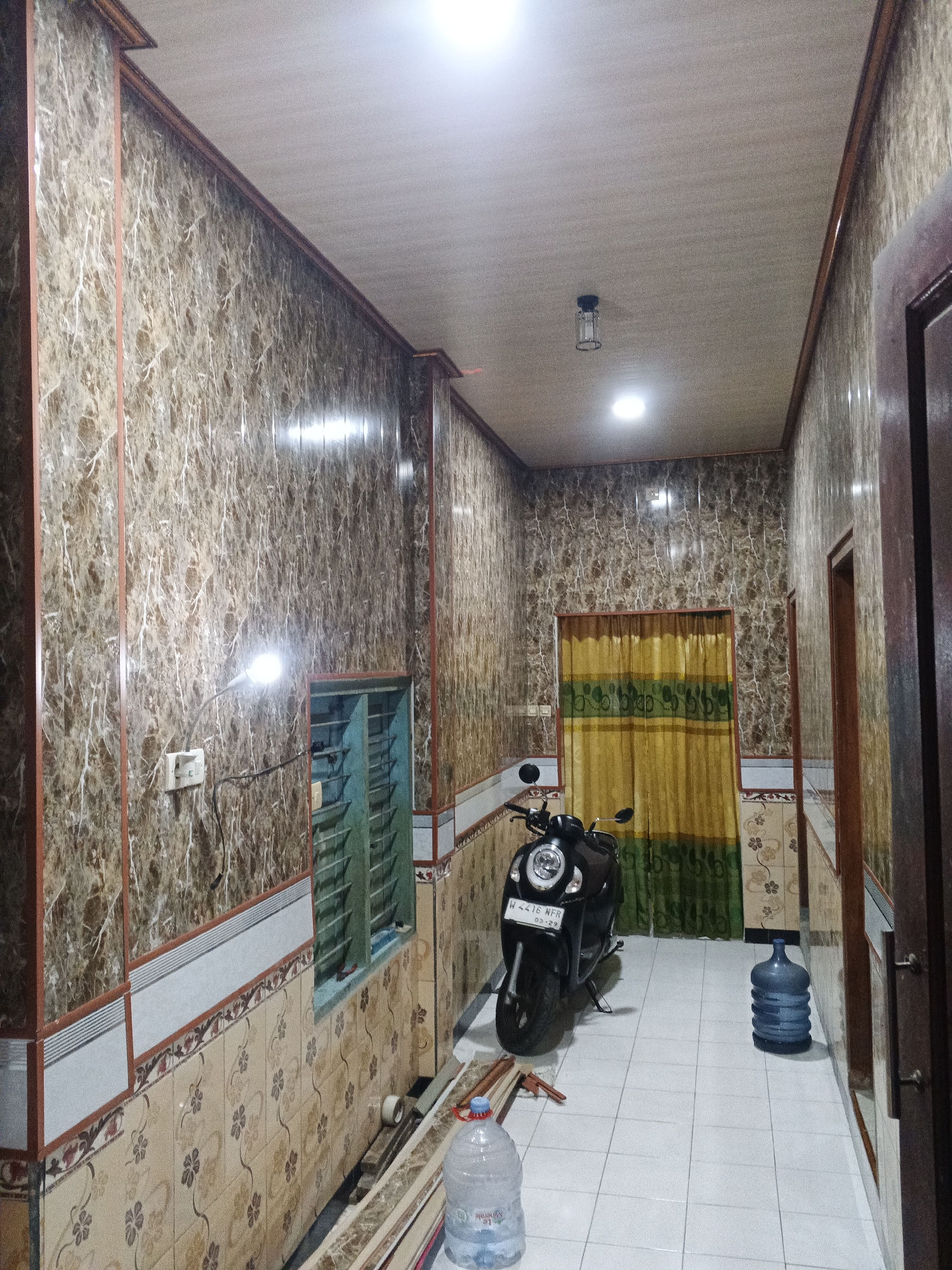 The image depicts an indoor hallway with ornate marble-patterned walls and a tiled floor. On the left, a motorcycle is parked, and on the right, a large water cooler jug is visible. The hallway is illuminated by ceiling lights and a floor lamp. The overall impression is one of a residential or commercial space. The scene is located in Kemangsen, Indonesia.