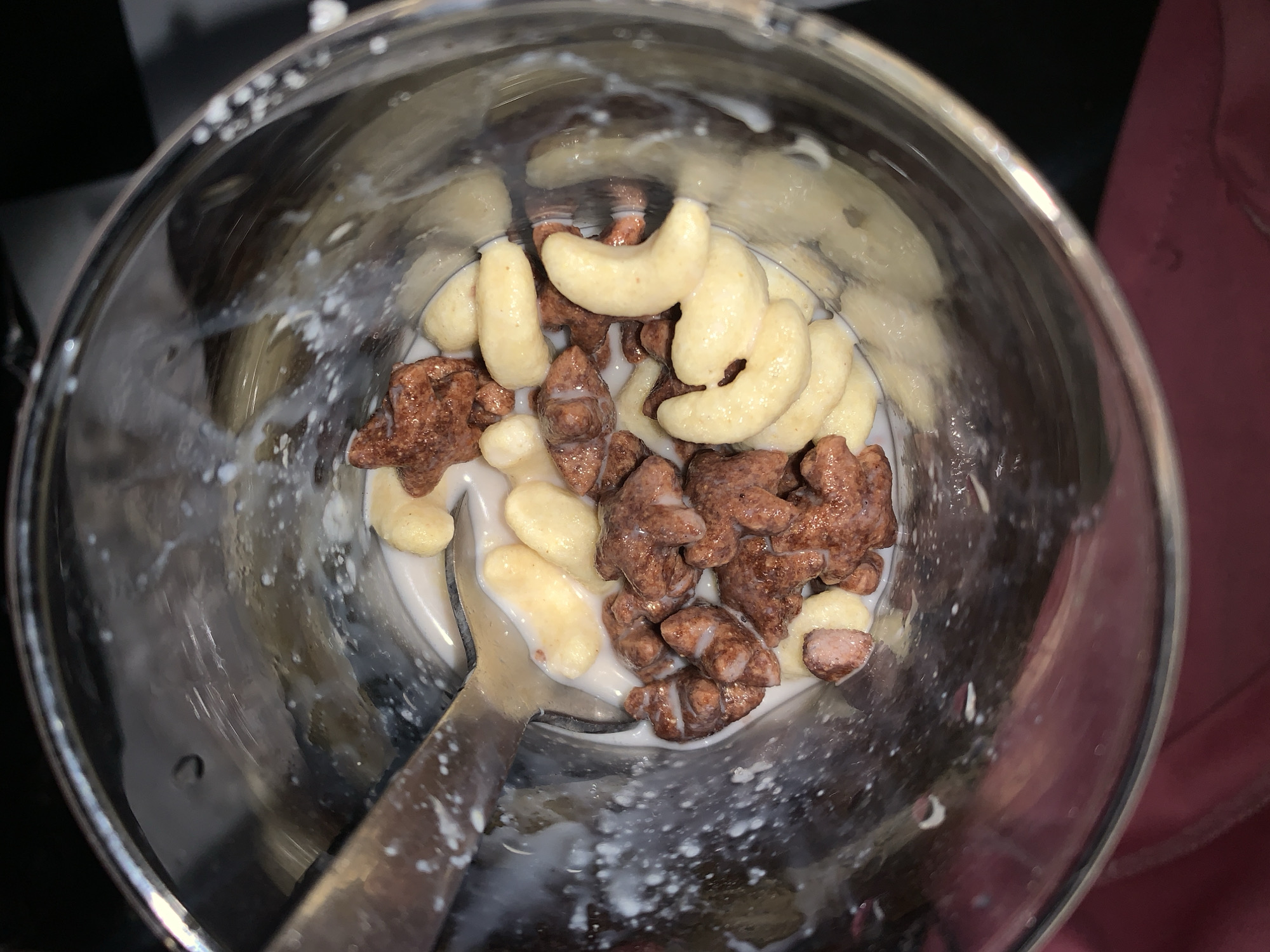 A top-down, close-up shot reveals cereal and milk in a clear bowl. The cereal consists of light-yellow, crescent-shaped pieces and dark-brown, bear-shaped pieces. A metal spoon rests in the bowl. Milk partially covers the cereal. The bowl sits on a dark surface, partially bordered by what appears to be a pink object. There is no indication of Rafin Par, Nigeria.