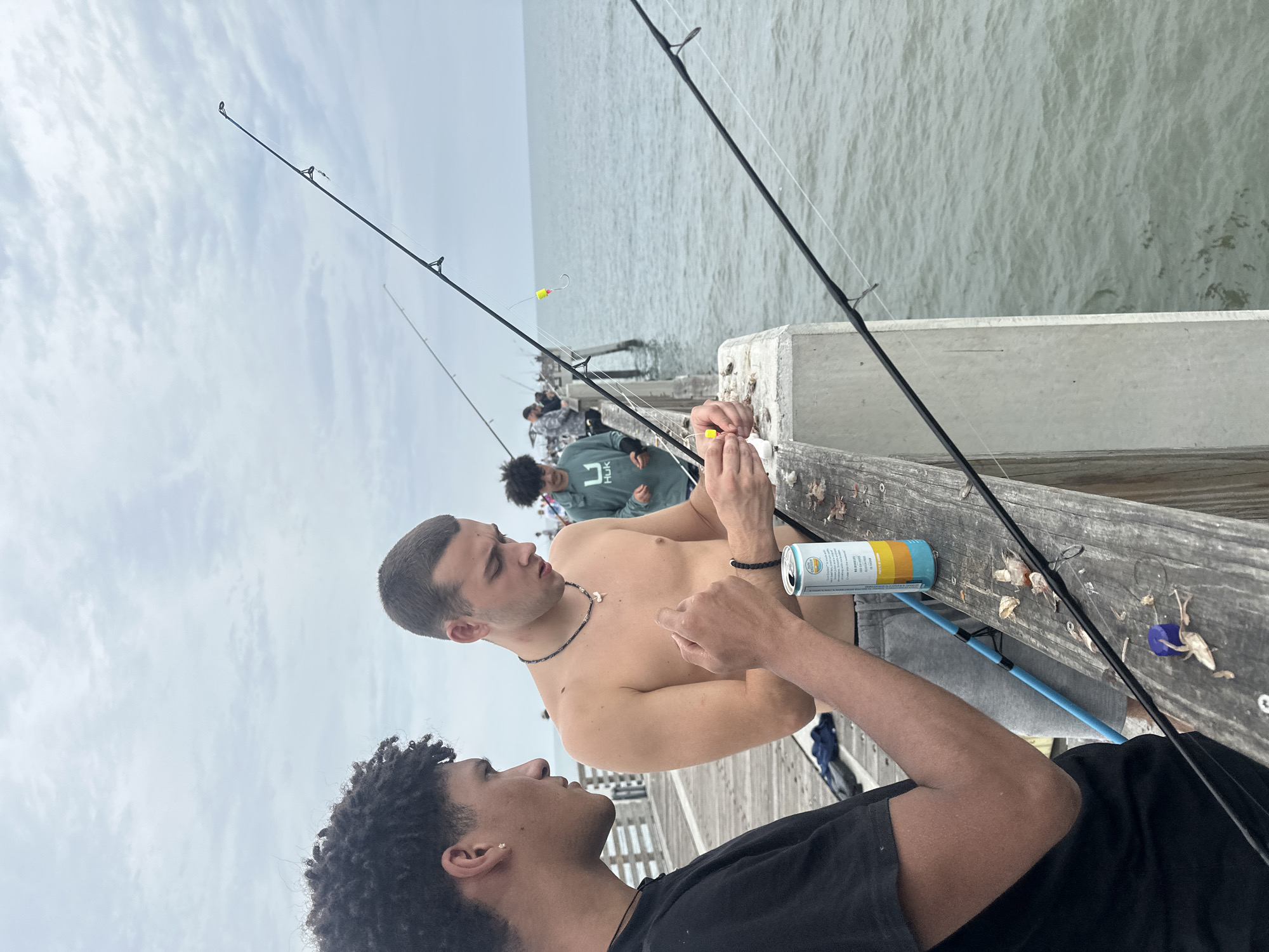 The image shows a group of people fishing on a pier. The pier appears to be in Jacksonville Beach, Florida. Two men are in the foreground, one shirtless and the other wearing a black t-shirt. A can of beer sits on the pier next to them. Further back, other individuals are fishing as well, against a backdrop of the ocean and sky. The day appears overcast.