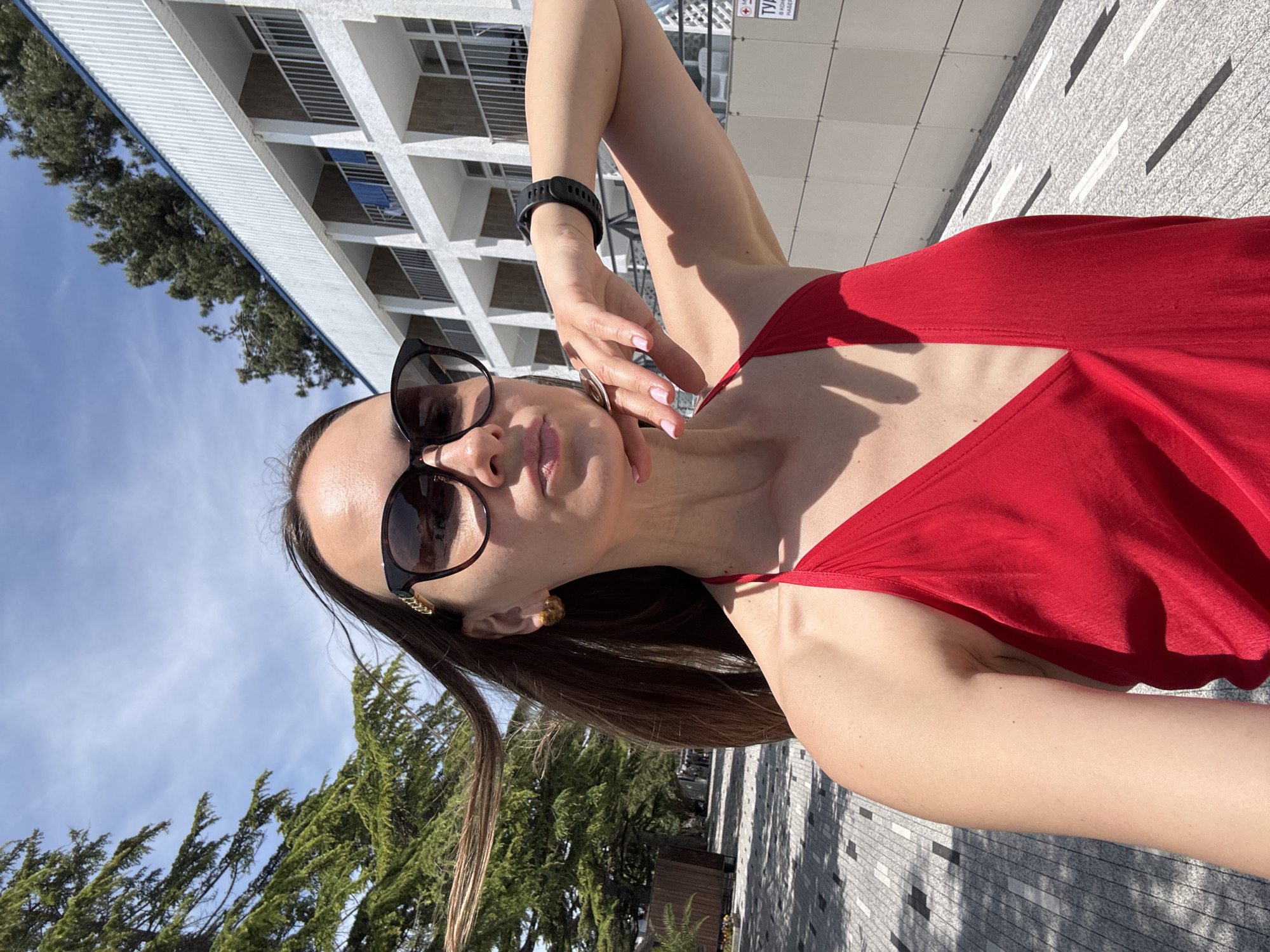 The image is a bright, sunlit selfie-style portrait of a woman standing outdoors. The woman occupies the lower and central portions of the frame, with her head and torso visible. She has long, dark brown hair, which is slightly uplifted by a breeze on her left side. She wears large, dark sunglasses featuring a gold-toned chain-link detail on the temples. Her lips are slightly parted in a pout. She is dressed in a vibrant red, V-neck, sleeveless top, possibly made of satin or a similar smooth fabric. Her left hand is raised to her chin and neck, with fingers lightly touching her skin, and her fingernails are painted a light pink. A black digital smartwatch is visible on her right wrist.
The background to the woman's left features a paved pedestrian pathway composed of square and rectangular blocks, exhibiting patterns of sunlight and shadow cast by overhead trees. Large, dark green coniferous trees, possibly cedars or pines, are prominent on the left side of the frame and extend upwards into the top left corner. In the background to her right, a multi-story white building with a series of identical balconies is visible. The building has dark-colored railings on the balconies and a blue band running along its upper facade or roofline. Additional coniferous trees are visible above and behind this building. The sky above is clear and bright blue with sparse, wispy white clouds. On the far right, affixed to the building, a partially visible white sign displays a red cross symbol and Cyrillic text, which includes the words "ТУАЛЕТ" (TOILET) and "НАБЕРЕЖНОЙ" (EMBANKMENT). The architectural style, vegetation, and public promenade setting with Cyrillic signage indicating an embankment are consistent with a location within the city of Sochi, Russia.