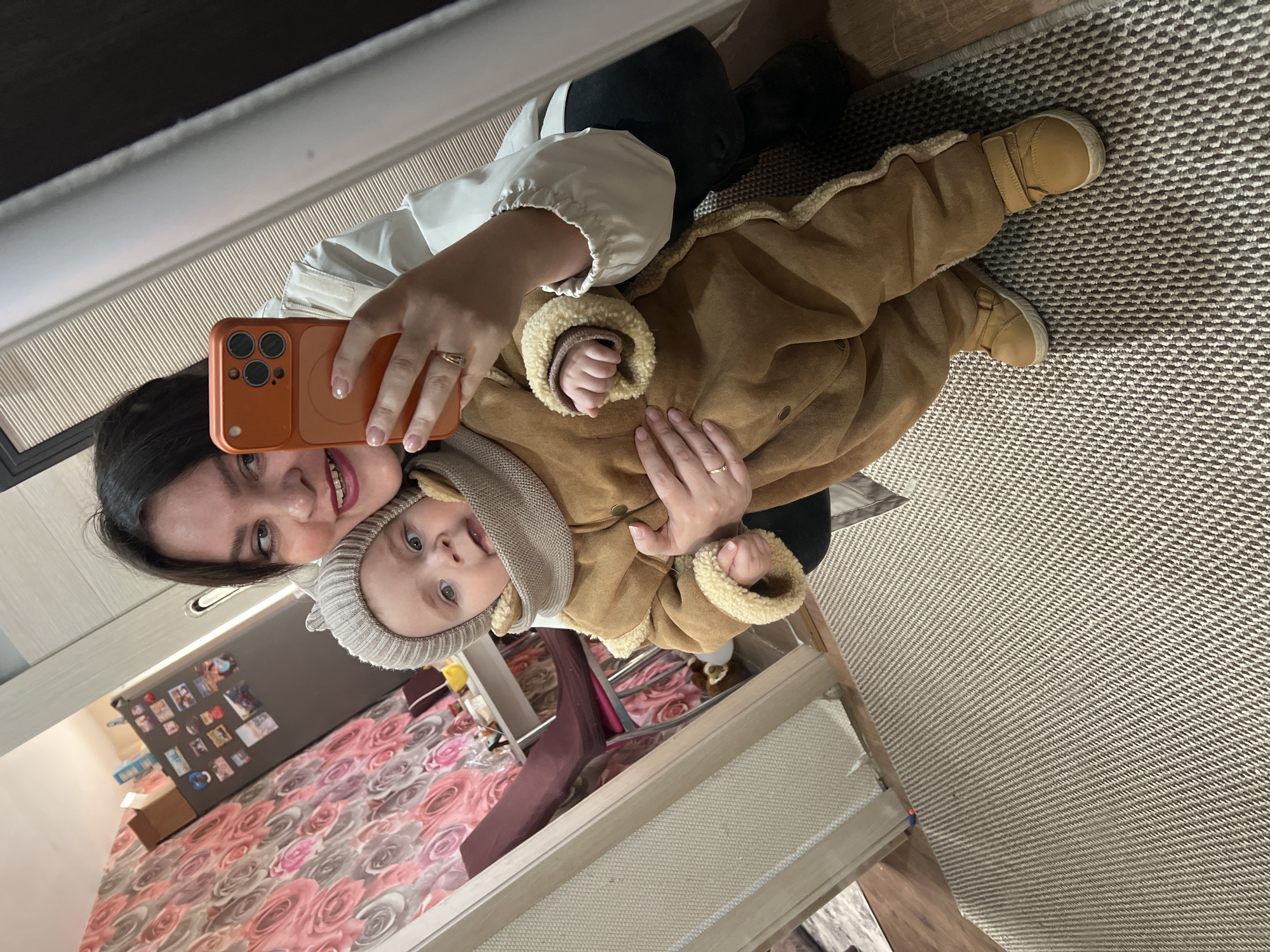 A woman is holding an infant, wearing a brown coat and hat, for a selfie. The phone being used is orange and has three rear-facing cameras. The woman has dark hair and is smiling. The background includes a patterned rug, part of a bed with a floral bedspread, and a cabinet or dresser. There is also what looks like a refrigerator covered in photographs. The location is likely inside a home. The city mentioned, "Noviy, Russia," is not discernible from the visual data.