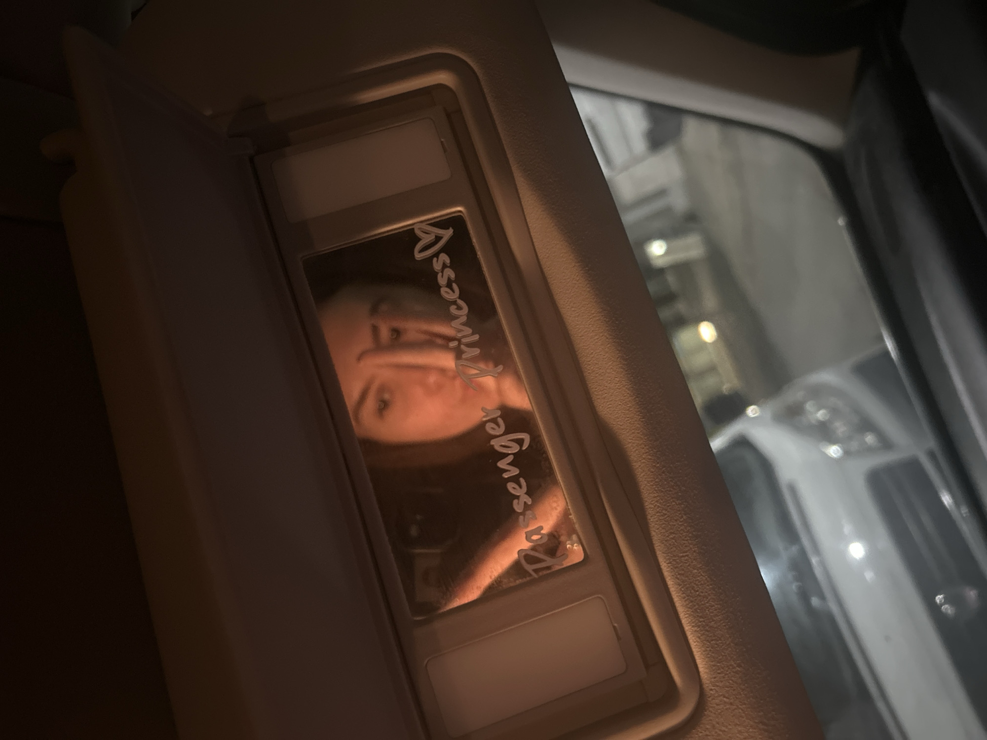 The photograph presents an interior view of a vehicle, focusing on the passenger-side sun visor. The visor is in a lowered position, revealing a mirror. Reflected in the mirror is a person, partially obscured, with the words "Passenger Princess" written on the mirror's surface. The background of the reflection indicates an outdoor setting with blurred lights and the silhouette of a vehicle. The overall lighting suggests it is either nighttime or a dimly lit environment.