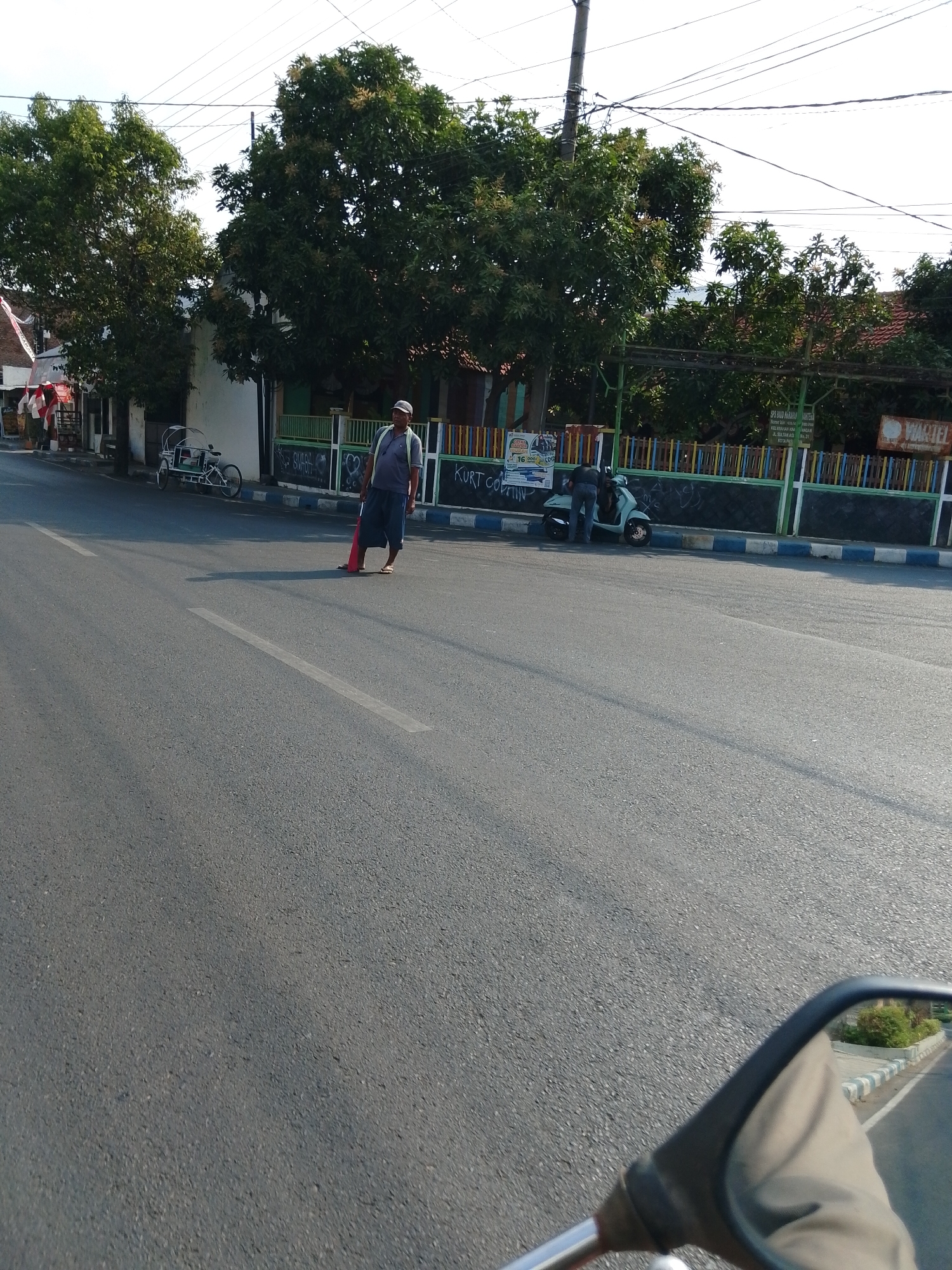 A man is standing on a road in Pasuruan, Indonesia, holding a red object. He is wearing a grey t-shirt, blue shorts, a cap, and sandals. The road is paved and appears to be a city street. There are trees and buildings in the background. A motorcycle is parked on the side of the road. The scene is captured from the perspective of someone riding a motorcycle, as indicated by the presence of a mirror in the foreground.