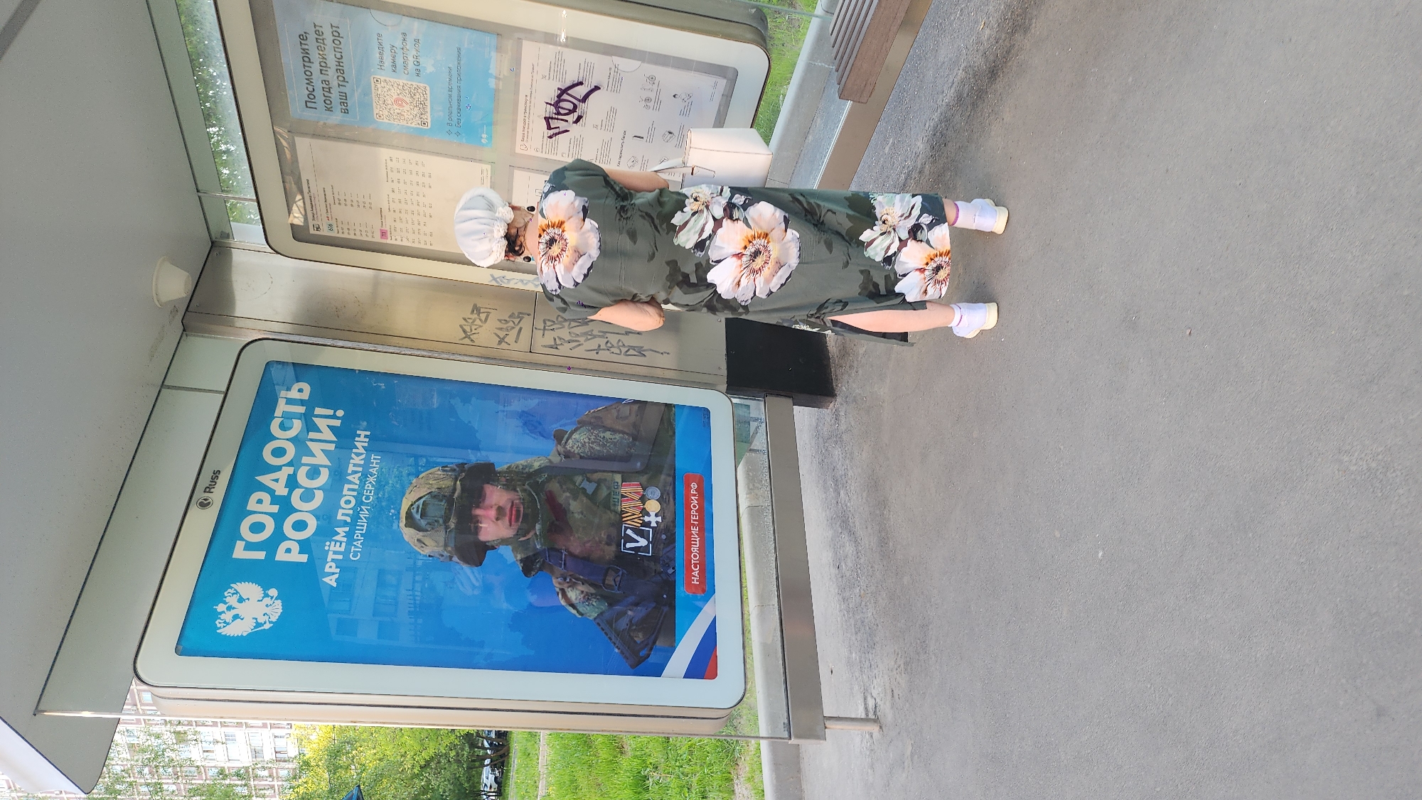 Here's a forensic description of the image:
**Setting:** The image was taken at a bus stop in Moscow, Russia.
**Objects:** A bus stop structure is visible, including an advertising panel featuring a Russian soldier, Artem Lopatkin, labeled as a "Hero of Russia." The advertisement includes a QR code for information.
**People:** An elderly woman with a white head covering and glasses is standing at the bus stop, next to the advertisement. She is wearing a floral dress and white tennis shoes with pink socks. She is carrying a white purse.
**Environment:** The surrounding environment includes green vegetation and buildings. The ground is paved.