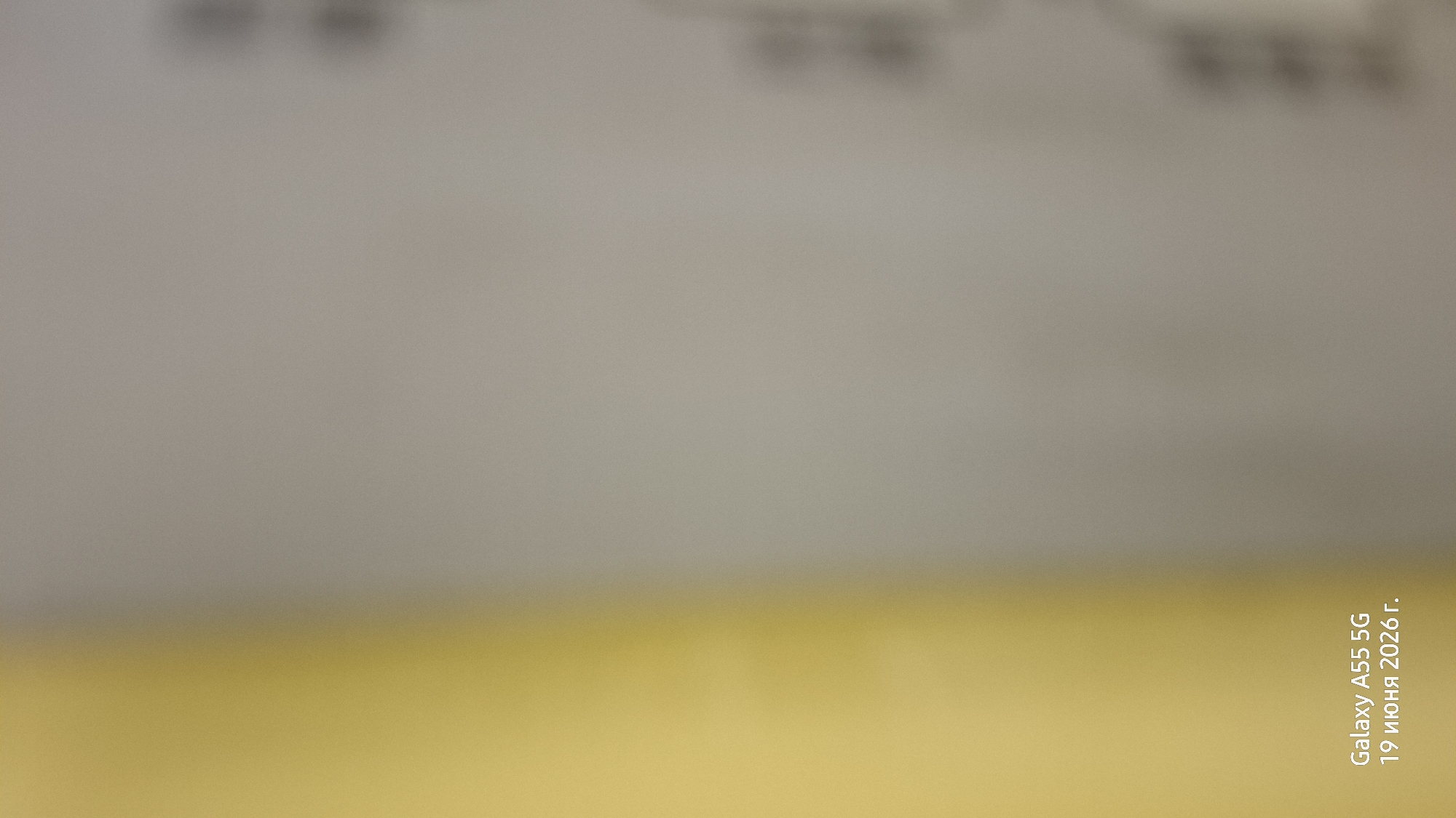 This is a media file of a document, likely a piece of paper, with some text printed on it. The document is out of focus and appears to be viewed from an angle. The top portion of the document is a light grey, and the bottom portion is a bright yellow. There is some blurred text or imagery at the very top of the grey section.
In the bottom right corner of the image, rotated vertically, is a timestamp or watermark. It reads "Galaxy A55 5G 19 июня 2026 г.". This indicates the photograph was taken with a Samsung Galaxy A55 5G phone on June 19, 2026, at 19:00. The date being in the future suggests this is either a placeholder or a pre-set timestamp. The "г." at the end of the date is a Russian abbreviation for "год" (year). The text "19 июня" translates to "June 19".
The overall impression is that of a document that has been captured with a mobile device, with the metadata embedded directly into the image. The lack of focus and the stark color contrast between the grey and yellow suggest it might be a form, a receipt, or some other printed material. No people or specific objects are clearly discernible. The setting appears to be indoors, given the lighting and the nature of the photographed item.